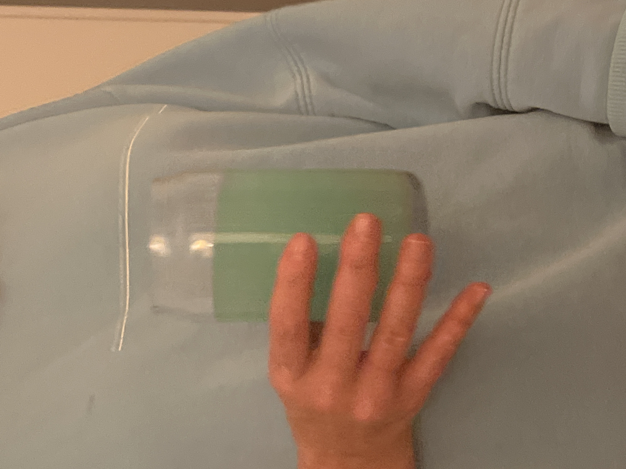 A photograph shows a fair-skinned hand covering a portion of a light blue garment, possibly a sweatshirt or similar item. The hand is positioned palm down, with the fingers extended. Partially visible beneath the hand, encased within what appears to be a transparent plastic cover, is a green object. The background consists of the light blue garment, with some textural details suggesting fabric folds or seams. The color palette is dominated by light blues and greens, with skin tones providing contrast.
The location is unknown.
