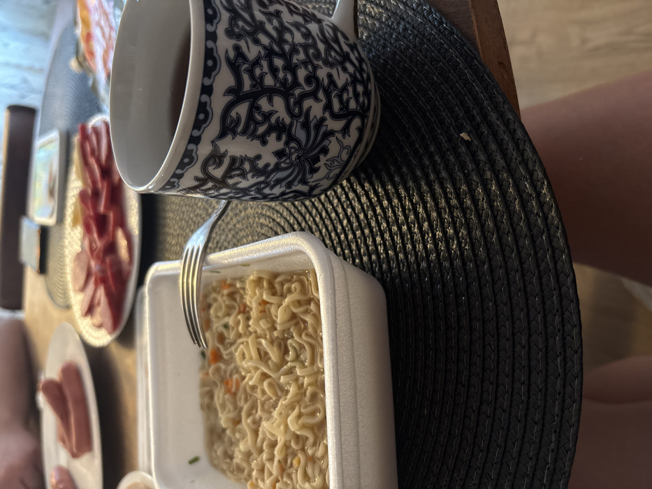 The image captures a close-up, high-angle view of a dining table in Lezhnevo, Russia, during a meal. In the foreground on the left, a white rectangular styrofoam container holds cooked instant noodles in broth, with visible orange and green specks. A silver fork rests horizontally across the rim of this container, with its tines partially immersed in the noodles. To the right, a white ceramic mug, decorated with intricate dark blue floral and foliate patterns, contains a dark liquid. Both the container and the mug are positioned on a circular, dark-colored woven placemat. The placemat rests on a wooden table surface, visible on the right. In the blurred background, a white plate on the left holds several cooked sausages, sliced lengthwise. Another white plate on the right displays multiple slices of cured meat, arranged in a circular pattern. A person's bare leg is partially visible in the lower right corner, and a blurry limb is discernible in the upper left background, indicating the presence of individuals.