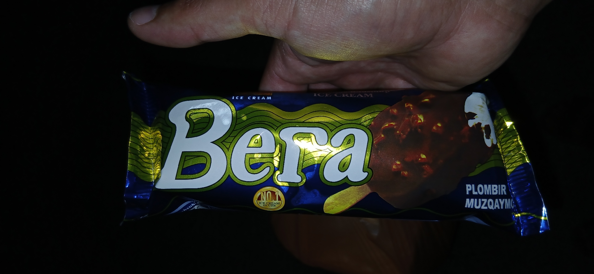 A hand is holding a wrapper for a chocolate-covered ice cream bar. The wrapper is predominantly blue with wavy yellow and green accents. Large white text on the wrapper reads "BEIZA" vertically. A circular emblem with "NO1" in yellow on a red background is visible to the left of the "B". Below the name, text reads "ICE CREAM". Further down the wrapper, the words "PLOMBER MUZOQAYMO" are visible in white text on a blue background, indicating the flavor. The ice cream bar itself, visible through the wrapper, appears to be chocolate-coated with embedded nuts or chocolate pieces. The image is taken in a dimly lit environment. There is no information in the image to indicate the location in Andijani, Uzbekistan.