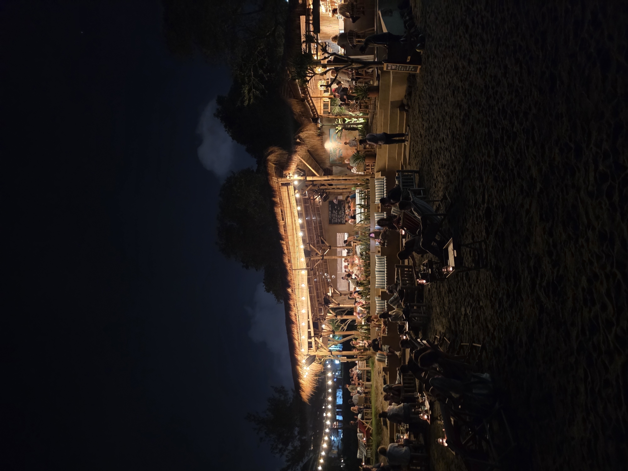 At night, the beachside restaurant in North Kuta, Indonesia, is filled with patrons. The restaurant has a thatched roof and is illuminated by string lights. People are seated at tables, some eating and drinking. The beach is dark.