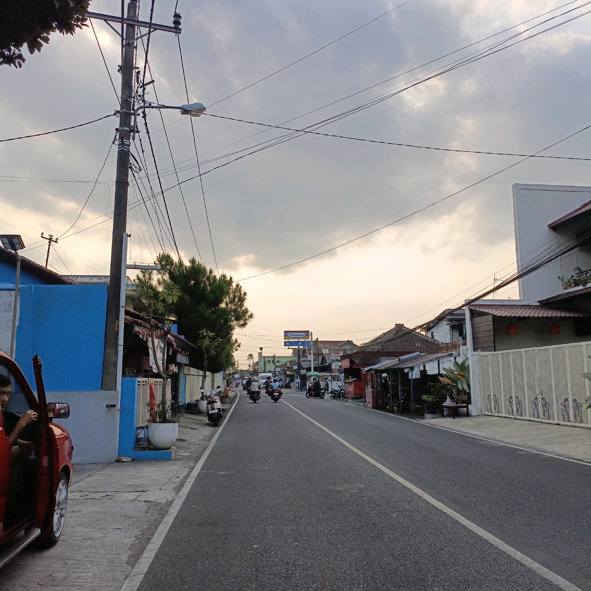 A street scene in Salatiga, Indonesia, features a paved road with a white lane marker, flanked by buildings and trees. The sky above is overcast with a warm, soft light, suggesting either dawn or dusk. On the left side of the road, a red car is parked, its door open, with a person sitting inside, looking towards the left. Adjacent to the car is a blue building with white trim. Further down the street, on both sides, are various establishments, including shops with awnings and signage, some appearing to be food stalls. Several motorcycles are visible, both parked and in motion, with people riding them. The overall impression is of a moderately busy urban street during a transitional time of day.