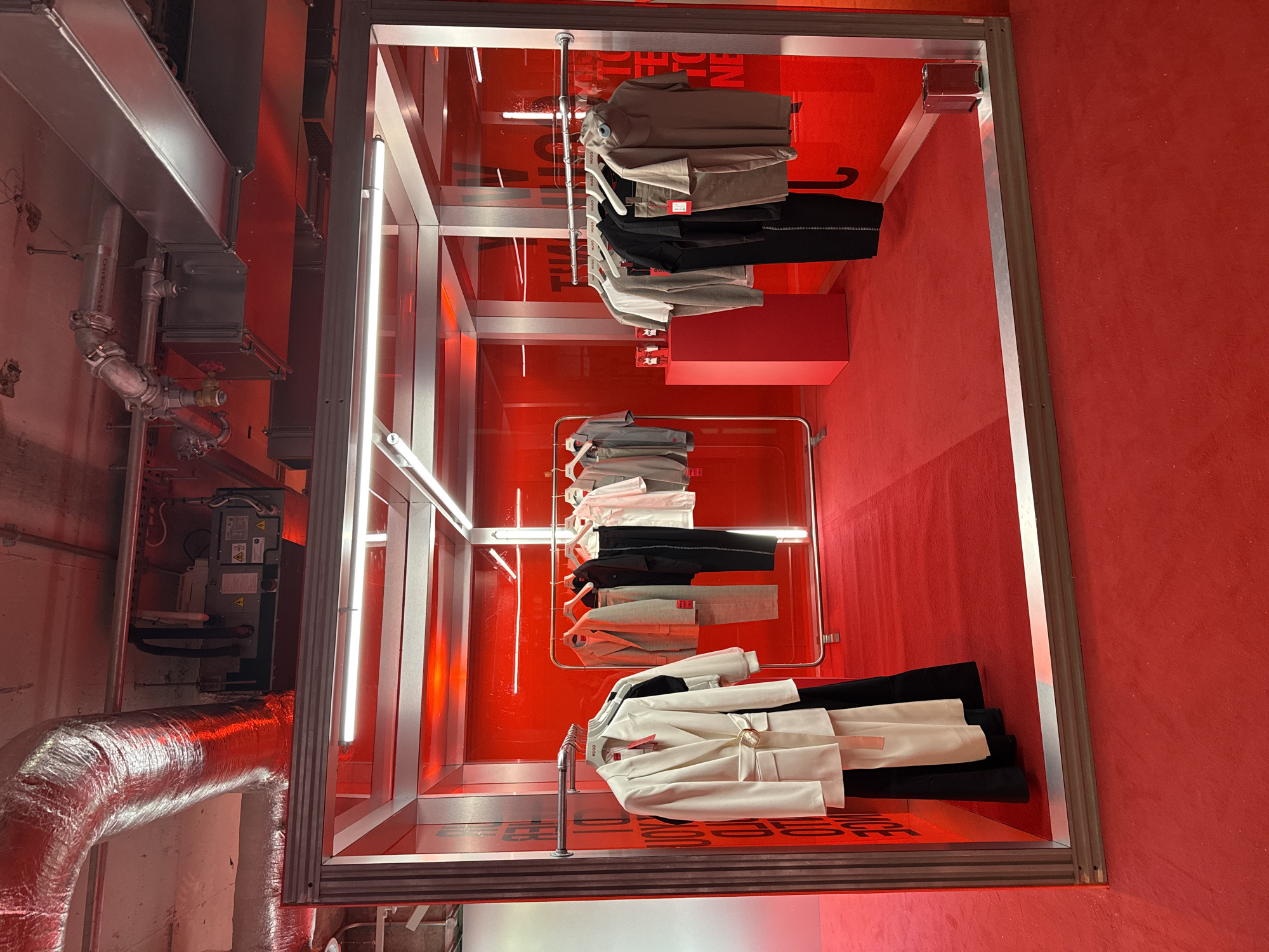 The image shows an interior space, possibly a retail environment or exhibition space in London, United Kingdom, judging by the general aesthetic. The dominant colors are red and silver/grey. The walls and floor are bright red, contrasting with the metallic silver framing and fixtures.
Three vertical displays of clothing are arranged in a linear fashion. Garments, including shirts, trousers, and jackets, are hung on racks within each display. The clothing is predominantly in neutral colors such as white, black, grey, and beige.
Mechanical and industrial elements are visible on the left side of the image, including exposed ductwork and piping. The overall impression is of a space that is modern and minimalist.