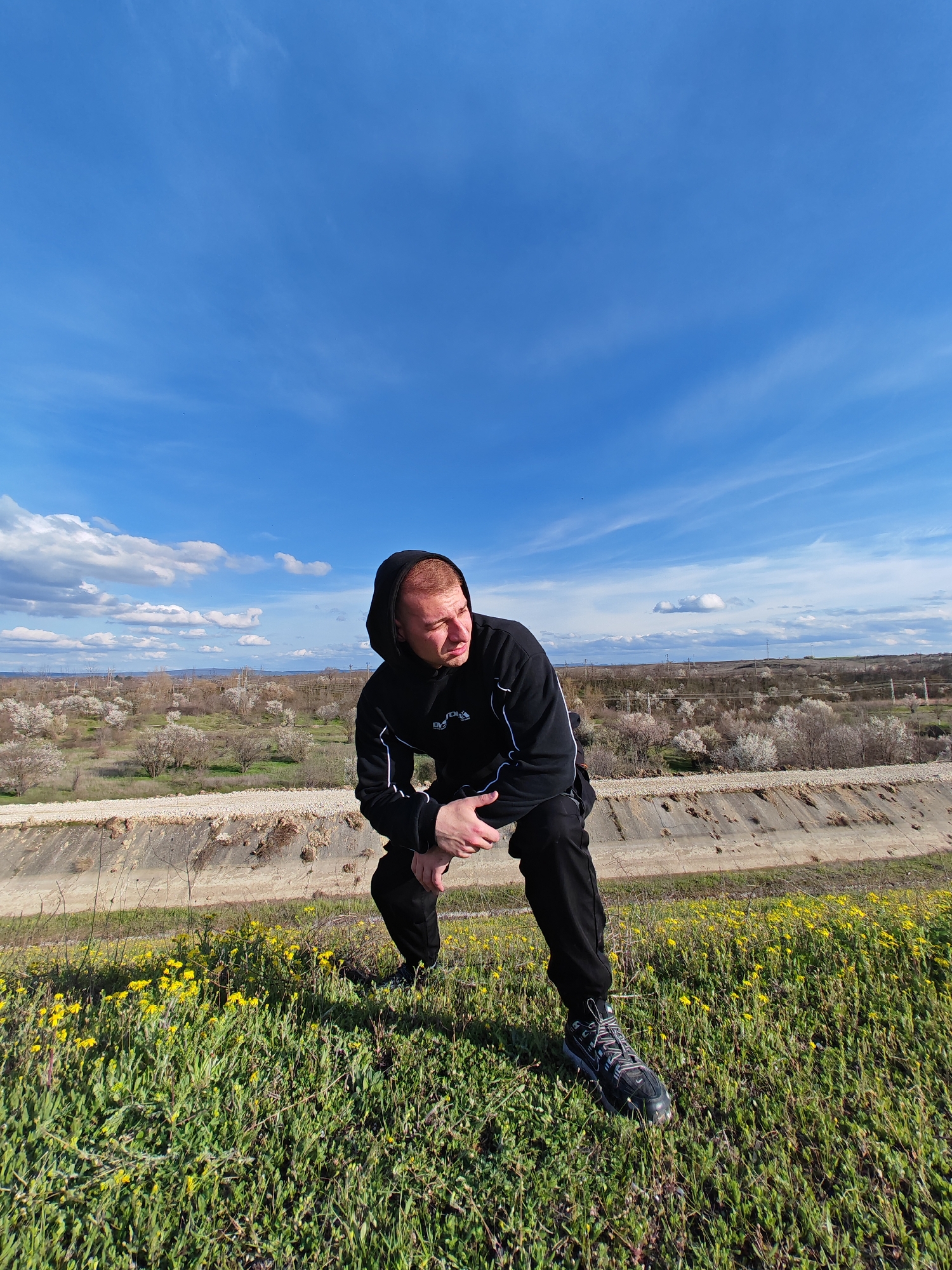 A man wearing a black hoodie and black pants is crouching on a grassy hill with yellow flowers. He is looking to his right. Behind him, there is a concrete canal and a field of trees with white blossoms. In the background, there are buildings and power lines. The sky is blue with scattered white clouds. The location is in Shumen, Bulgaria.