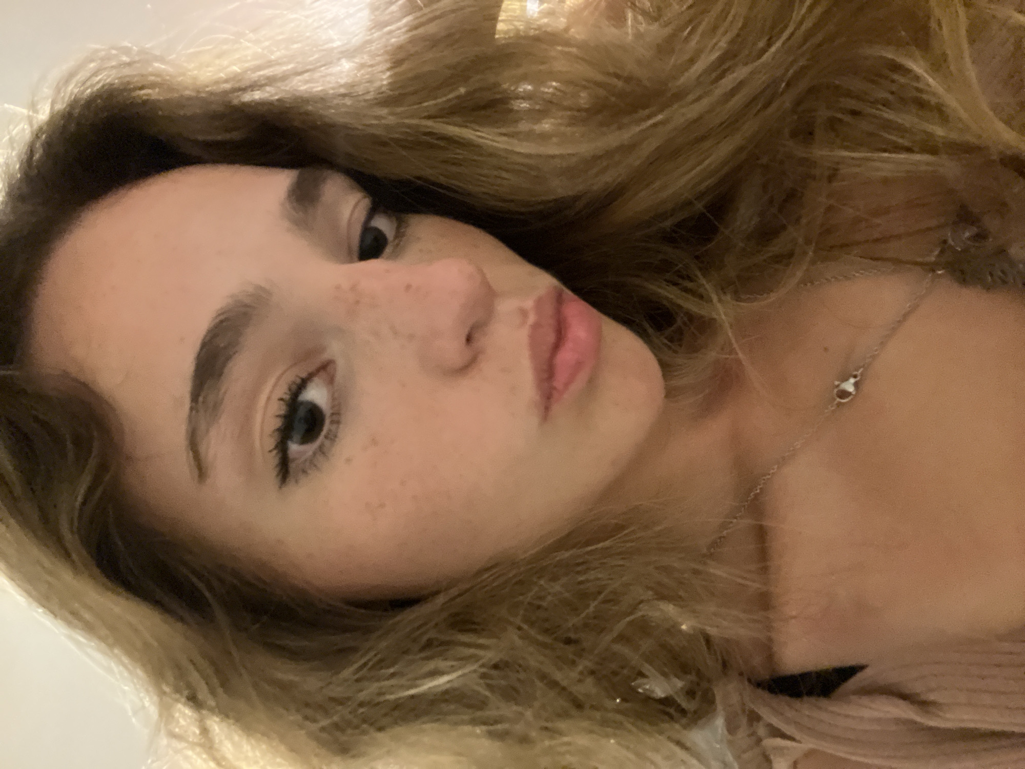 A close-up, angled selfie shows a young woman with fair skin, freckles, and light brown hair. Her left eye is clearly visible, with dark lashes and eyebrow. She has a small nose and pink lips, pursed slightly. A silver necklace with a pendant is visible at her neck. A light-colored garment is visible at the bottom of the frame. The background is out of focus. The city and country are not discernible from the image.
