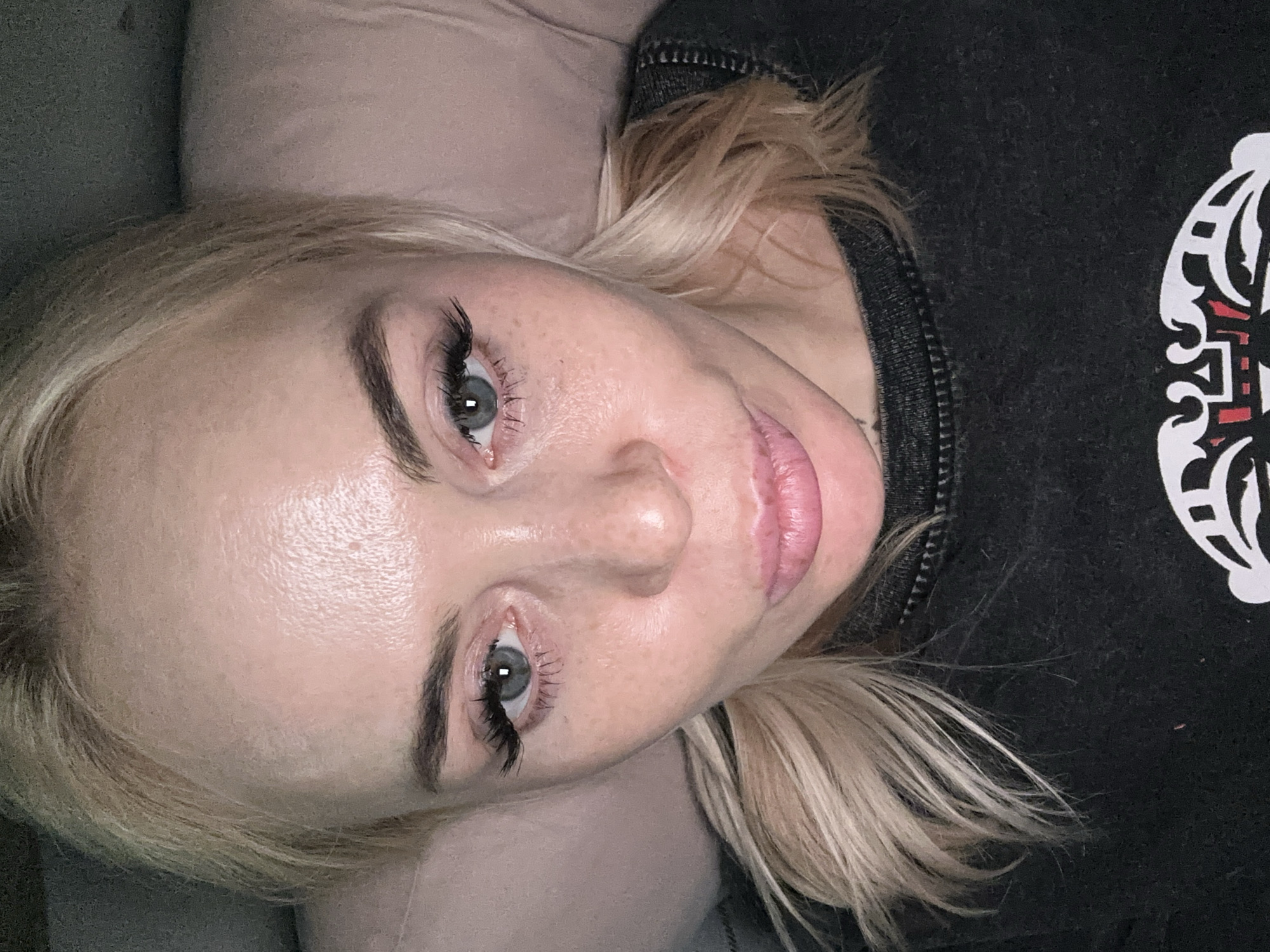 The original image captures a close-up, slightly tilted view of a person, likely female, from the chest up. She has fair skin, blonde hair, and light blue or grey eyes. Her hair is casually styled, framing her face. She is wearing a dark-colored sweater or top, which appears to be black or dark grey, with a white and red patterned design visible on the upper right side of the garment. Her eyelashes appear long and full, possibly indicating extensions or mascara, and her eyebrows are well-defined. Her lips are slightly parted, revealing a natural pink hue. The person is looking directly into the camera with a neutral to slightly serene expression. Behind her head, a light, soft surface, possibly a pillow or bedding, is visible, suggesting an indoor setting like a bedroom or living room. The lighting appears soft, illuminating her face. There is no visible text in the image. The provided cropped images offer closer views of her eye, skin texture, hair, and a part of her sweater design.