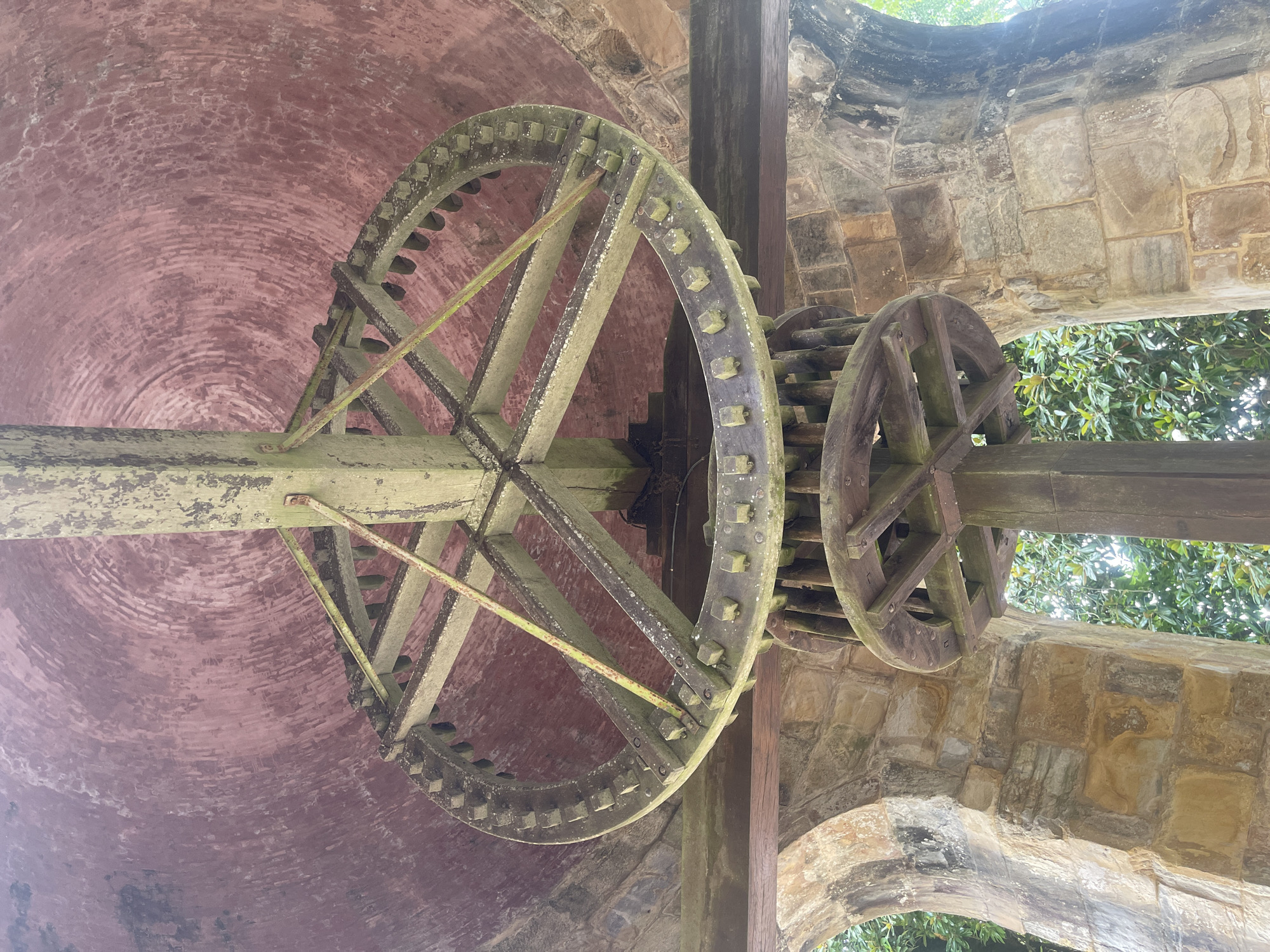 This image shows an interior view of a mill mechanism within the gardens of the Mafra National Palace in Mafra, Portugal. The central focus is a large, wooden gear wheel, likely part of a water or wind-powered system. The wheel has a toothed outer rim and is supported by wooden spokes radiating from a central axis. Below this larger wheel, a second, smaller wooden wheel with a similar spoked structure is visible.
The mechanism is housed within a circular structure, suggested by the domed, reddish-pink ceiling made of bricks, evident at the top of the frame. The walls appear to be constructed from rough-hewn stone blocks, forming arched openings through which greenery and trees are visible, indicating an outdoor setting within the palace grounds. The wooden components of the mill show signs of age and weathering, with moss or lichen growth and chipping paint on some surfaces. The lighting suggests it is daytime, with natural light illuminating the scene. There are no people or animals present.