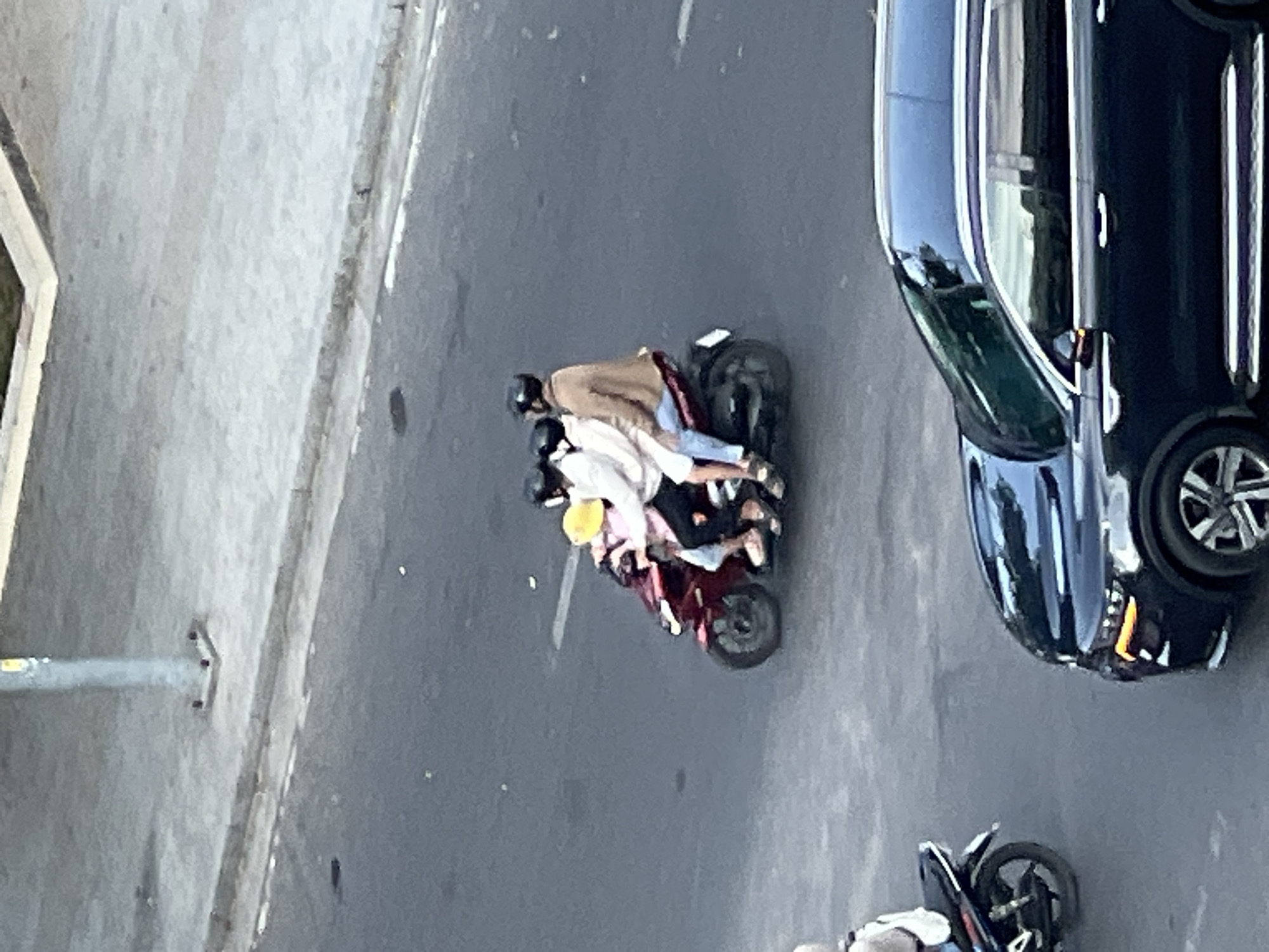On a paved road in Nha Trang, Vietnam, four people ride a single red motorcycle. The rider and three passengers, all wearing helmets, are positioned in a staggered arrangement, with the driver at the front. The driver wears a brown jacket and blue jeans, while the passengers are dressed in lighter colored clothing and dark pants. A yellow helmet is visible on one of the passengers. The motorcycle has a license plate that is not legible. To the right, a dark-colored SUV is partially visible on the road. In the background, a gray concrete sidewalk with a light pole and curb is visible. The scene suggests a typical urban street in Vietnam.
