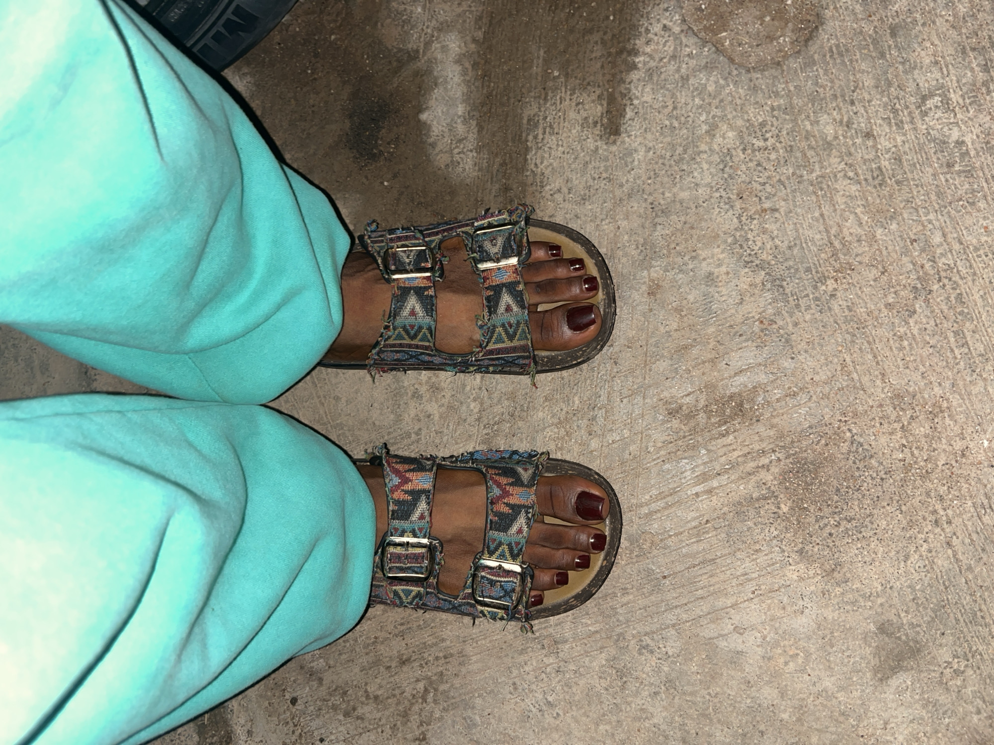 A close-up, eye-level shot shows a person's feet wearing turquoise sweatpants and patterned Birkenstock-style sandals standing on a concrete surface. The sandals have two buckled straps with a colorful, geometric fabric pattern and a brown cork footbed. The person's toenails are painted a dark red. The concrete ground appears weathered with visible lines and some scattered dirt or small stones. The overall scene is dimly lit, suggesting an indoor or evening setting. There is no indication of the specific location in Jos, Nigeria, within the image itself.