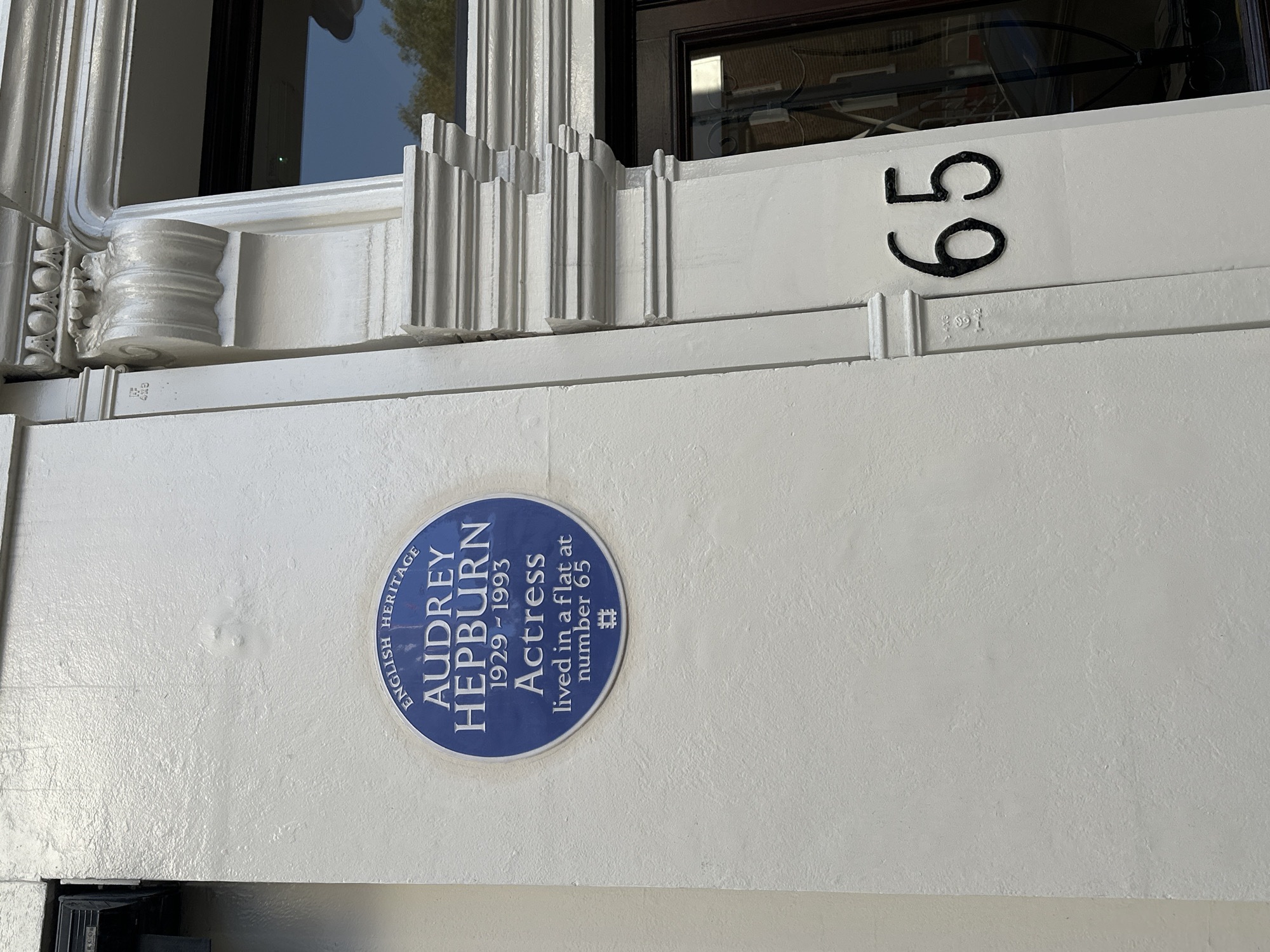The image shows the façade of a building in London, United Kingdom, featuring a blue plaque honoring actress Audrey Hepburn, who lived in a flat at number 65. The plaque is mounted on a white wall, with the address number "65" displayed in black numerals above it. Architectural details such as moldings and a windowsill are visible.
