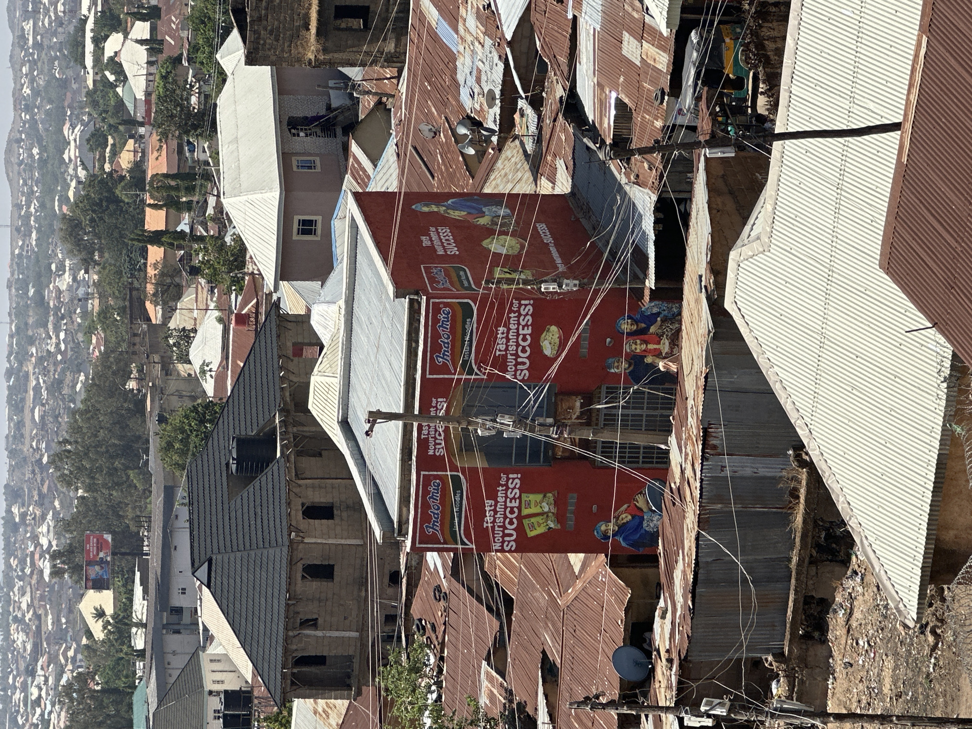 The image is an elevated view over a densely populated urban area in Jos, Nigeria, characterized by a patchwork of corrugated metal rooftops and a mix of informal and structured housing. The scene is dominated by a prominent red sign advertising "Indomie Instant Noodles," highlighting the text "Tasty Nourishment for SUCCESS!" The signs are affixed to buildings amidst a network of electrical wires and utility poles. In the background, a hazy horizon shows the extent of the urban sprawl against a backdrop of distant hills. The building construction materials suggest a mix of economic conditions within the area.