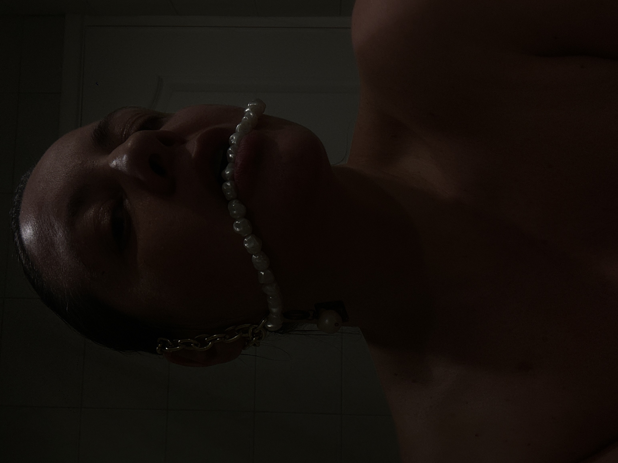 A close-up, low-angle photograph presents a person with their head tilted back, mouth slightly open, and a pearl necklace strung across their lips. The person appears to be partially nude, with visible skin on their chest and neck. A gold chain is also visible near the ear. The background consists of a dimly lit tiled wall, suggestive of a bathroom setting. The location is not identifiable as Riga, Latvia from the visual information provided.