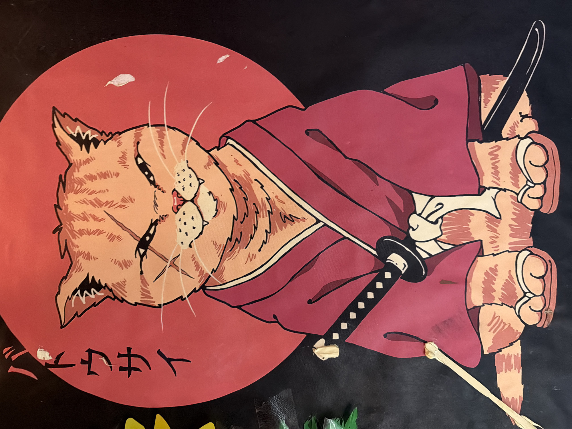 The image depicts a cartoon-style illustration of an orange tabby cat dressed as a samurai warrior. The cat is wearing a red robe with a white collar, and a black katana sword is visible, either held by the cat or attached to its attire. A large red circle is behind the cat, reminiscent of the Japanese flag. Japanese characters are inscribed below the cat in the circle. The illustration is on a black background, and the image includes parts of other objects in the foreground, including yellow and green objects.
There is no evidence of the location in the city of Bat Yam, Israel in the image.