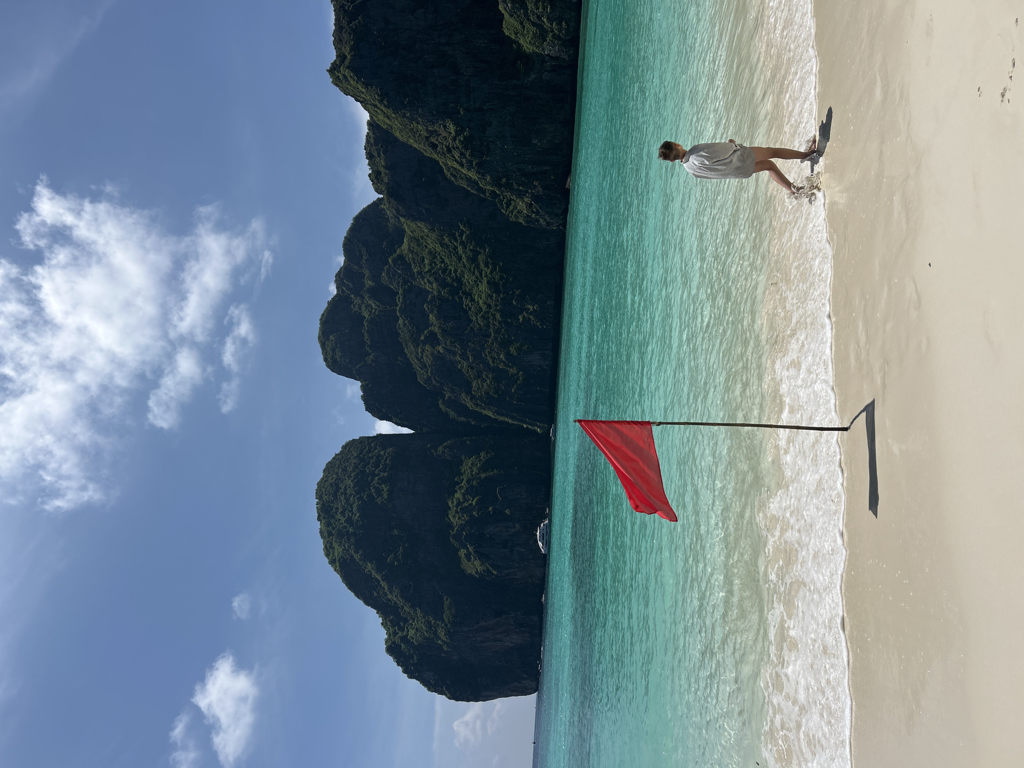 A lone person walks along a sandy beach beside clear turquoise water. In the foreground, a red flag on a pole waves in the breeze. Towering, green, rocky cliffs rise from the sea in the background under a bright blue sky with scattered clouds. A white boat is visible in the distance. The scene depicts a tropical beach setting in Ao Nang, Thailand.