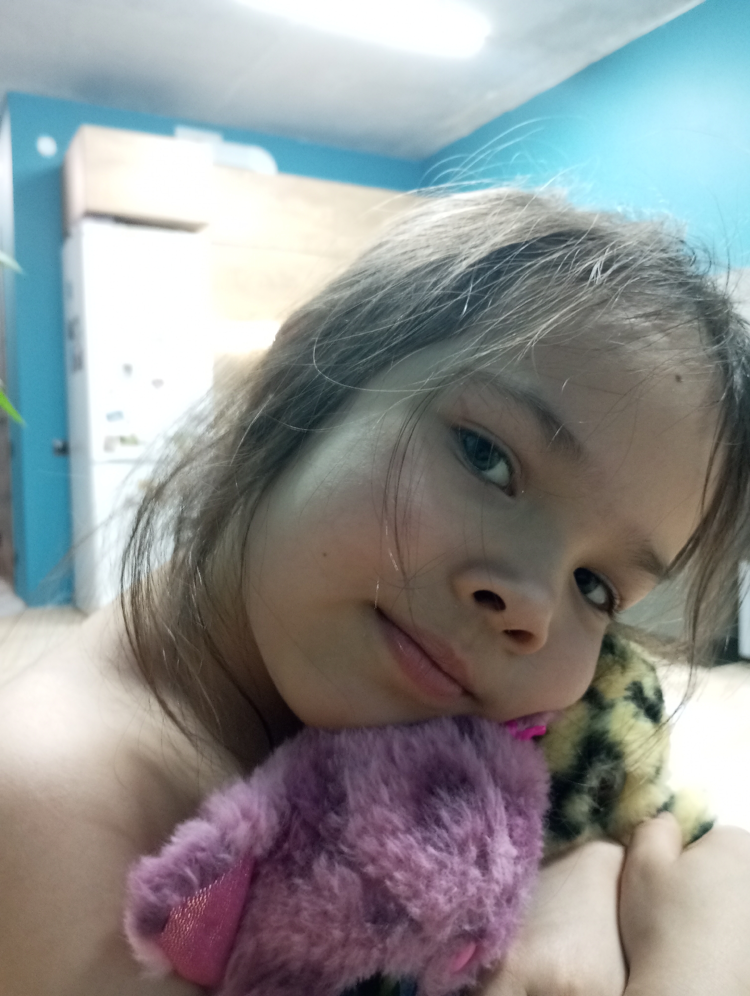 A young girl with brown hair and blue eyes is holding two stuffed animals, one purple and one yellow-spotted, close to her face. The background shows a blue wall and part of a white refrigerator, suggesting the location is indoors, possibly a kitchen. The location in the city of Pervomaysky, Russia cannot be verified or dismissed.