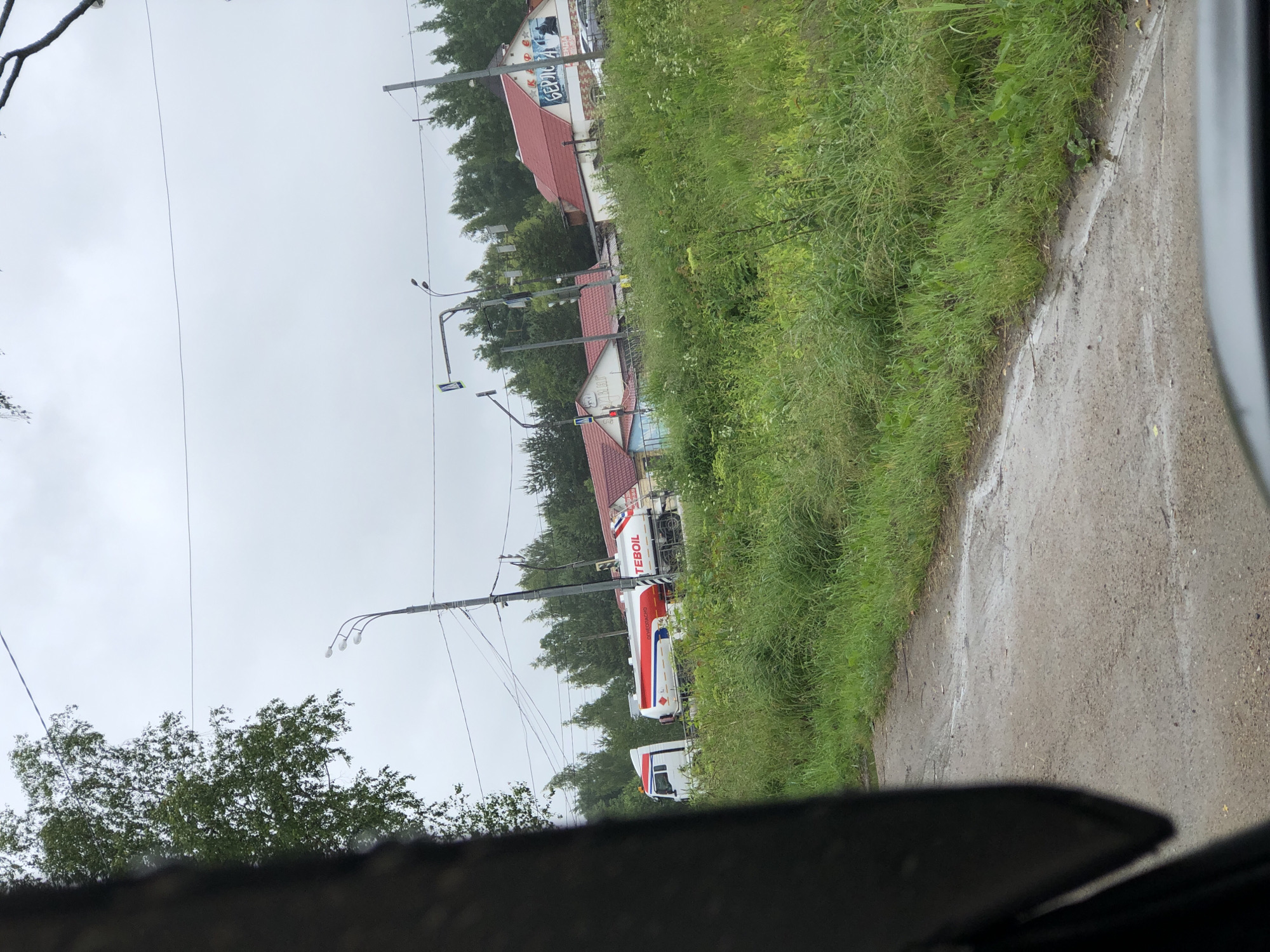 The image depicts a street scene in Safonovo, Russia, on a cloudy day. In the foreground, a portion of a dark vehicle's interior is visible, with the driver's side window framing the outdoor environment. The road surface appears wet, with puddles and a general dampness suggesting recent rain. Lush green grass and vegetation line the side of the road.
Further down the road, several buildings with red tiled roofs are visible. One building prominently features a large sign with text that includes "ТЕБОИЛ" and "БЕРЮЛ". A white tanker truck with red and white detailing is parked or moving on the road. Traffic lights and lampposts are present, indicating an urban or semi-urban setting. Trees and foliage are abundant in the background, forming a natural backdrop to the buildings and infrastructure. Several cars are parked near the buildings. The sky is overcast, contributing to the muted lighting of the scene.