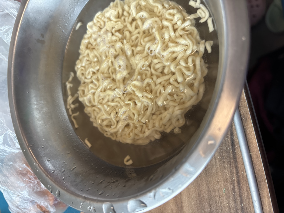 This image depicts a close-up, elevated view of an unidentifiable interior location in Tatarsk, Russia, focusing on a food preparation scene. A polished, silver-toned metallic bowl, possibly stainless steel, occupies the central and lower-right portions of the frame. Inside the bowl, a compacted block of light yellow, wavy instant ramen noodles is submerged in clear, hot water. Small bubbles are visible on the water's surface, indicating the noodles are in the process of cooking or soaking. Water droplets adhere to the exterior rim of the bowl on the left side. The bowl rests on a brown wooden surface, possibly a countertop or table, characterized by visible grain patterns. In the upper left background, a portion of a translucent plastic bag containing several orange-colored items, resembling carrots, is visible. Adjacent to the plastic bag, a light blue or teal surface is present. No individuals are visible in the scene.