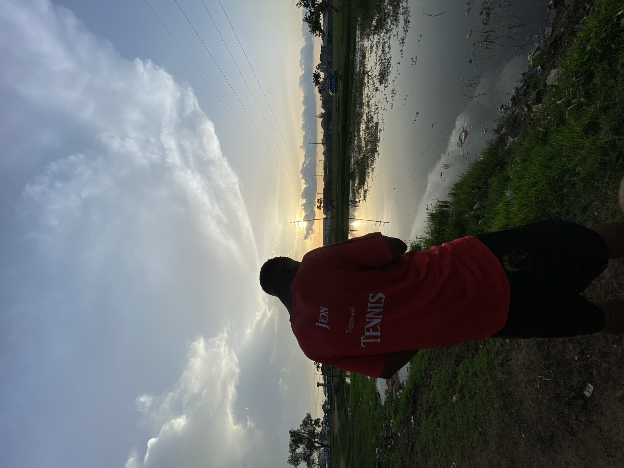 The image captures a sunset scene in Bukuru, Nigeria, featuring a lone individual from behind, a body of water, and a distant townscape. The central figure, a male, is seen in partial silhouette, positioned on the left side of the frame on a dirt path. He wears a red short-sleeved shirt with the words "JEON", "Brentwood", and "TENNIS" printed in white on the back. Black shorts with a light-colored emblem on the right rear pocket complete his attire. His head is tilted downwards, and his hands are held in front of him, obscured by the silhouette.
To the right of the individual and extending into the mid-ground is a still body of water, reflecting the orange, yellow, and blue hues of the sunset sky. The water's surface shows reflections of clouds and contains some floating debris. The immediate foreground features a mix of dirt and patches of green grass, with visible litter scattered along the path and near the water's edge.
The horizon reveals a flat landscape with numerous residential or commercial structures and sparse trees, indicating a populated area. The sun, a bright yellow-orange orb, is visible low on the left horizon, partially obscured by clouds and a series of parallel power lines stretching across the upper right portion of the sky. The sky above is characterized by scattered clouds, transitioning from bright, illuminated forms near the sunset to darker grey and blue higher up. A large, dark-leafed tree stands on the left side of the frame, behind the person.