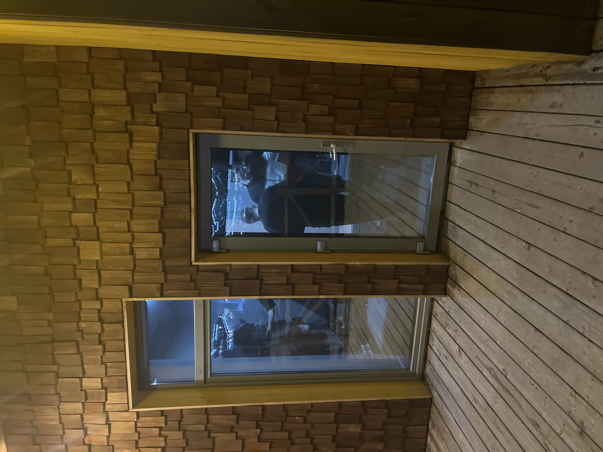 The image shows an exterior view of a building with two windows. The building is clad with light brown wooden blocks arranged in an irregular pattern. The windows have metal frames and are reflective, showing distorted images of people and lights inside. To the right of the windows, the wall is covered in light brown wooden planks running diagonally. The location appears to be Unknown, Unknown. There are no explicit actions, interactions or subjects to describe.