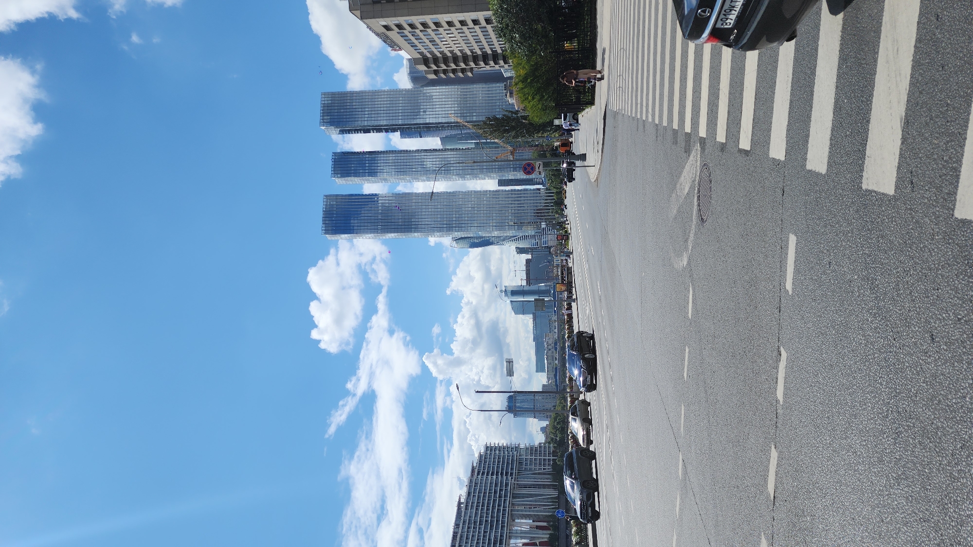 The image depicts a street scene in Moscow, Russia, likely in the Moscow City business district, characterized by modern skyscrapers and a wide asphalt road. In the foreground, a pedestrian crossing with white stripes is visible on the right side of the frame. A black car is partially visible on the far right, its rear bumper and license plate showing "B919KT". The road has clear lane markings, with dashed white lines dividing lanes. Several cars are driving on the road, including a dark SUV and a lighter colored sedan in the middle distance.
Dominating the background are several contemporary glass skyscrapers with blue-tinted facades. To the left, a long, multi-story building with a white facade and balconies stretches across the frame. The sky is blue with scattered white cumulus clouds, and the sun casts light, creating some lens flare in the upper left corner. A few figures are seen walking on the sidewalk and crossing the street in the distance, and construction cranes are also visible among the buildings, suggesting ongoing development. The environment appears to be an urban setting on a clear, sunny day.
