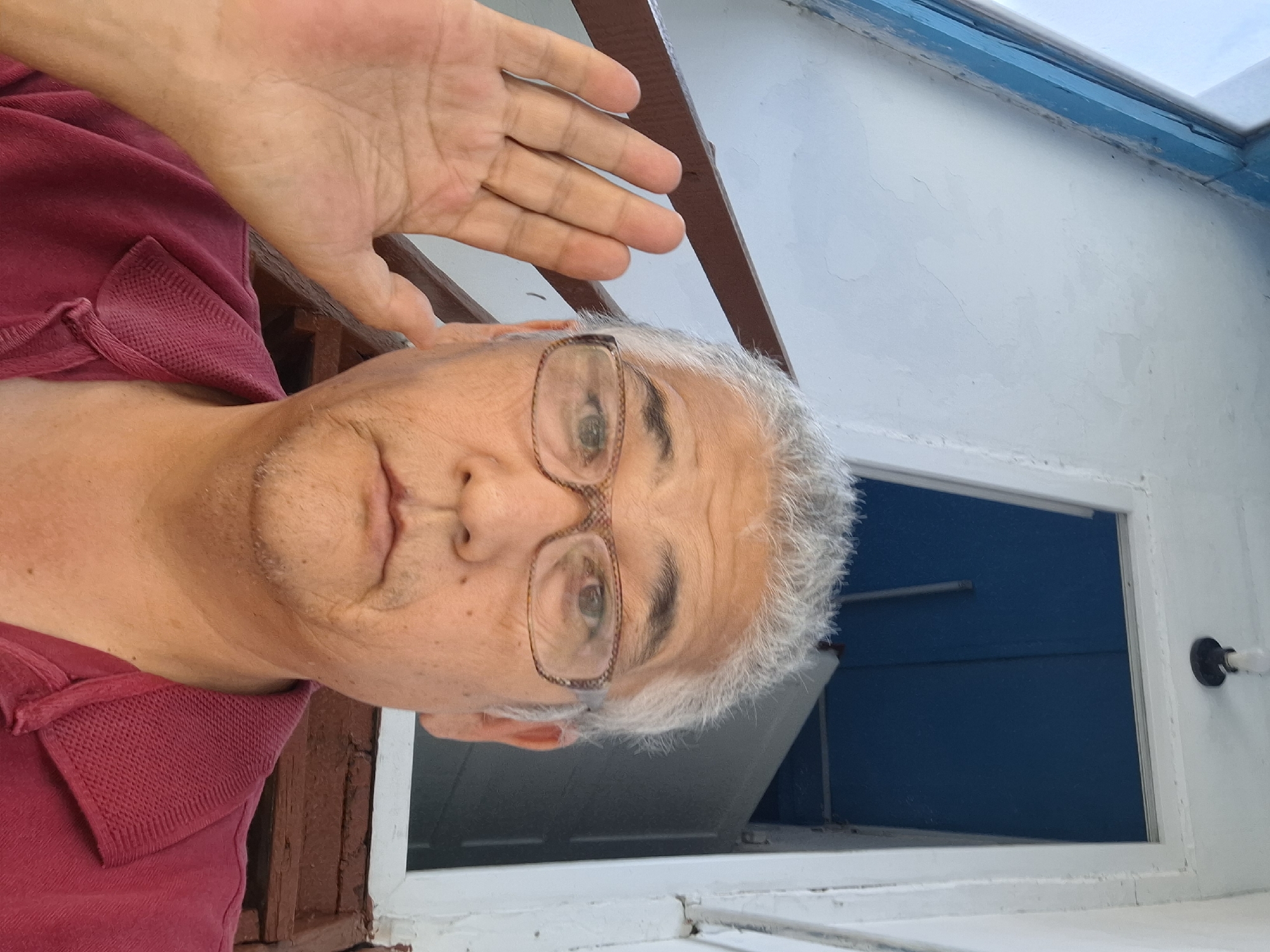 A close-up selfie shows an older East Asian man with grey hair and glasses, wearing a red polo shirt. He is holding his left hand up, palm facing the camera, with his fingers spread. The background suggests an outdoor setting with wooden stairs and a white-painted structure with a blue doorway. The image quality is decent, with good lighting and focus on the subject. There are no visible landmarks or clues that could pinpoint a specific location within Almaty, Kazakhstan.
