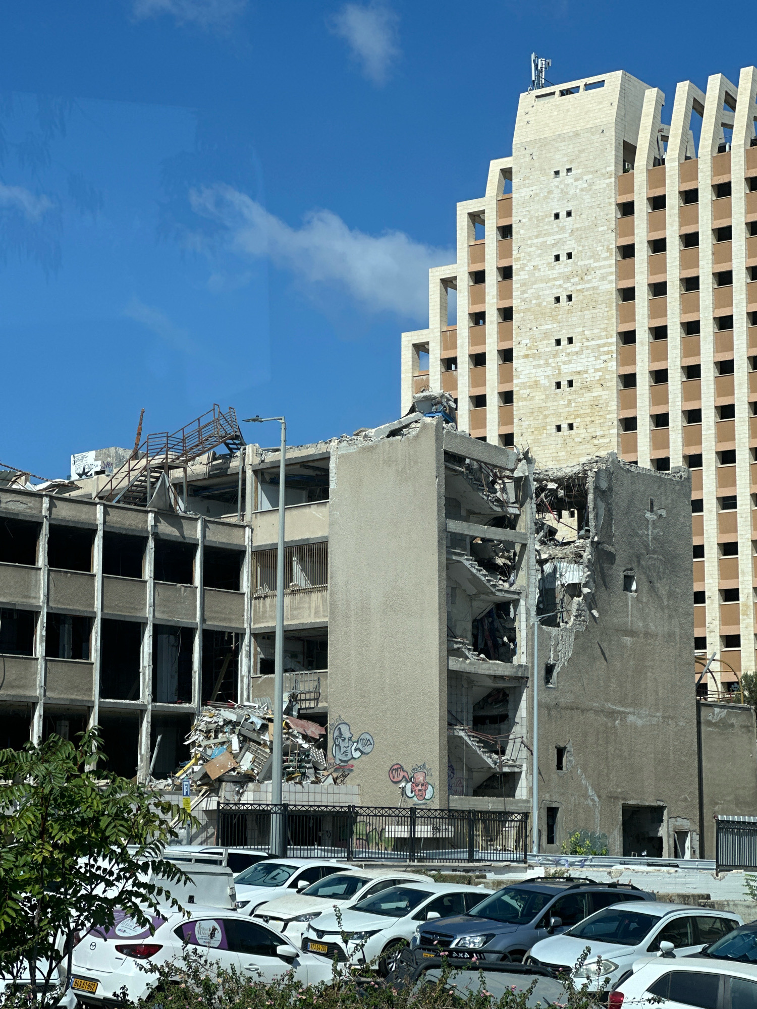 In an urban environment, a partially demolished concrete building stands in the foreground under a clear blue sky. The building is in a state of severe disrepair, with gaping holes in its facade, exposing interior structural elements and debris. A rusty metal staircase is visible extending precariously from the upper levels. Graffiti adorns some of the remaining walls. In the background, a taller, modern building with a beige facade and repeating window patterns rises above the damaged structure. The ground level features a parking lot filled with several white cars, some of which have purple decals on their windows. A few green trees and bushes are visible on the left side of the image, with more foliage in the foreground. The scene suggests an ongoing demolition or reconstruction project in an urban setting, possibly in the city of Haifa, Israel, based on the license plates visible on some of the cars.