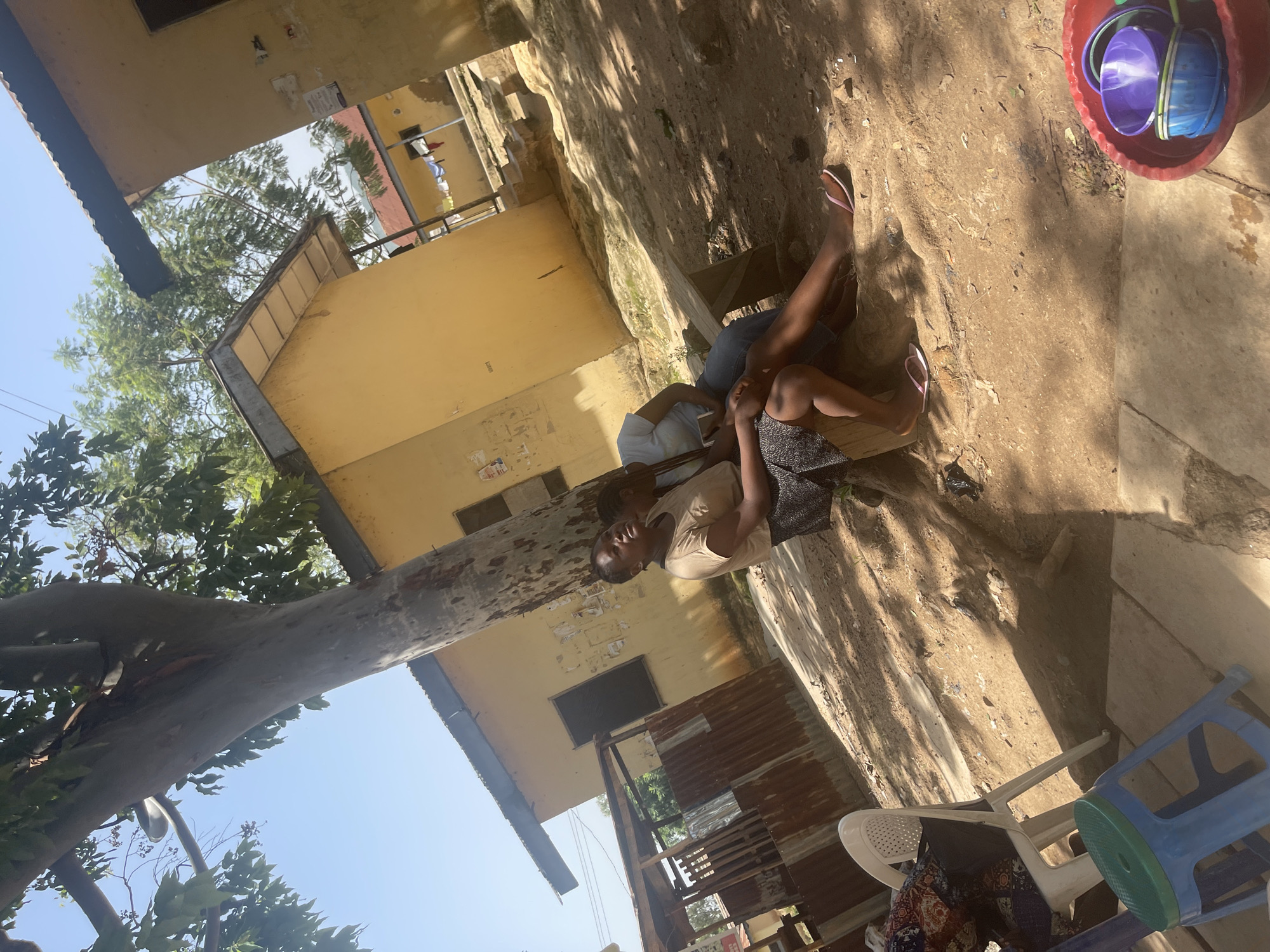 In Rafin Par, Nigeria, two women are seated on a wooden bench under a large tree. The woman on the left wears a tan shirt and a patterned skirt. The woman on the right wears a light blue t-shirt and blue jeans. Both are wearing flip-flops. Behind them are yellow buildings with corrugated metal roofs. The ground is dirt and scattered with debris. In the foreground, a white chair and a blue stool are visible, along with a red plastic basin filled with purple and green bowls. The scene is illuminated by bright sunlight, casting shadows.