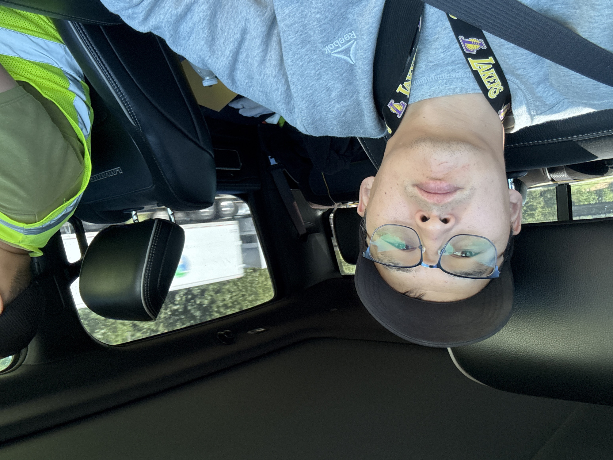 The image shows a man taking a selfie inside a vehicle. He is wearing glasses, a baseball cap, and a gray sweatshirt with a Reebok logo. A lanyard with a Los Angeles Lakers logo is around his neck. The car's interior is visible, including black leather seats and a sunroof. Outside the car, through the window, a highway with trees and a truck can be seen. Another person wearing a high-visibility vest is in the passenger seat. The location is identified as Ridgefield, United States of America.