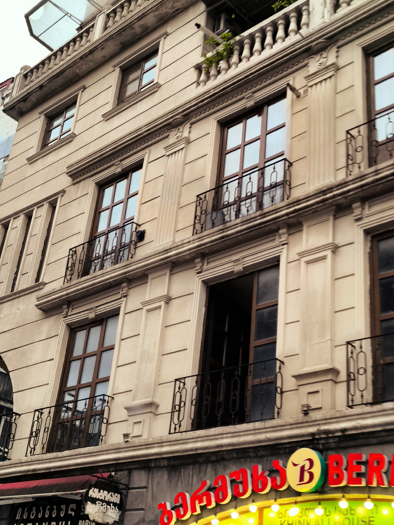 The image shows a multi-story building in Batumi, Georgia, with a cream-colored facade and classical architectural details. Several windows with brown frames and balconies with ornate metal railings are visible. The building appears to be of a commercial nature, as evidenced by signage at the bottom of the image. One sign, in a stylized font, displays "BER" in red letters, and another smaller sign features a circular logo with a "B" inside. Below this, there are smaller signs with text in what appears to be Georgian script, and an English phrase "KHINKALI HOUSE" is visible. The overall impression is that of a well-maintained building in an urban setting, possibly housing a restaurant or shop.