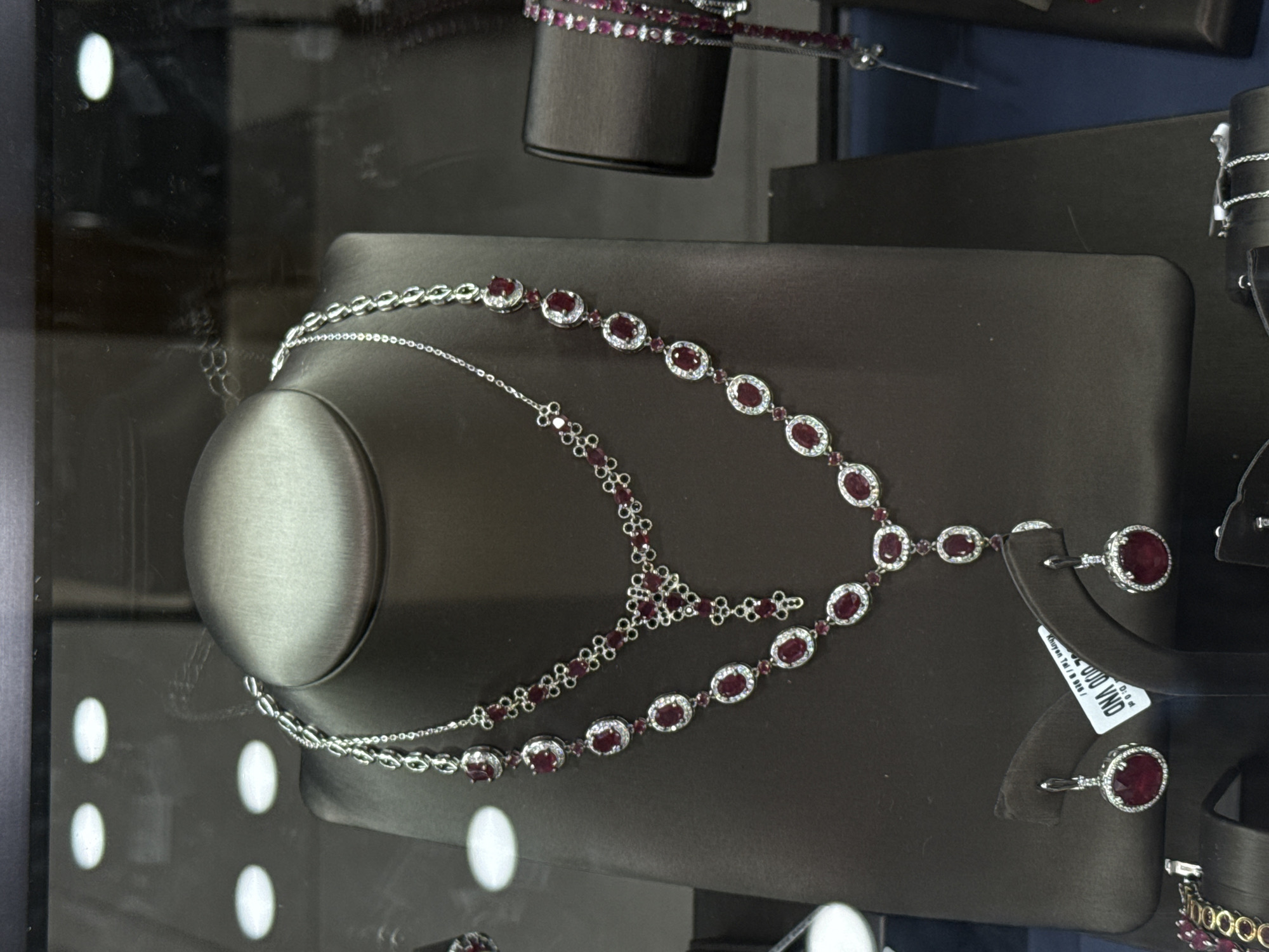The image shows a jewelry display inside a store located in Nha Trang Ward, Vietnam. A dark brown jewelry stand is on display containing a two-strand silver necklace with oval ruby stones. The necklaces feature silver chains. At the bottom of the jewelry stand are a pair of ruby earrings. A price tag is visible, displaying the price in Vietnamese Dong. Other jewelry displays are visible in the background. The reflections on the glass of the jewelry display show bright, white lights.