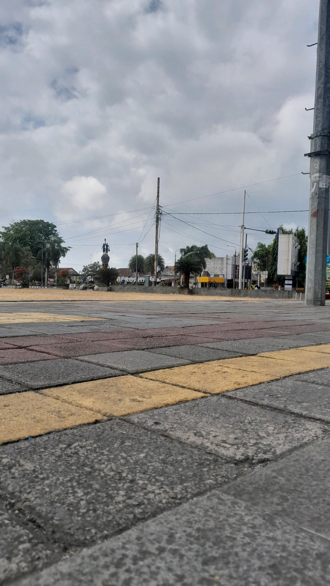 The image captures a low-angle view of a paved public square in Cilacap, Indonesia. The ground is covered in interlocking rectangular pavers in shades of gray, maroon, and yellow. In the background, a monument with three figures standing on a pedestal is visible, flanked by trees and palm trees. Beyond the monument, a street scene unfolds with buildings, traffic lights, utility poles, and electrical wires against a cloudy sky. A large, grey utility pole with visible brackets and tape is prominent on the right side of the frame. The overall impression is of a somewhat overcast day in an urban environment.