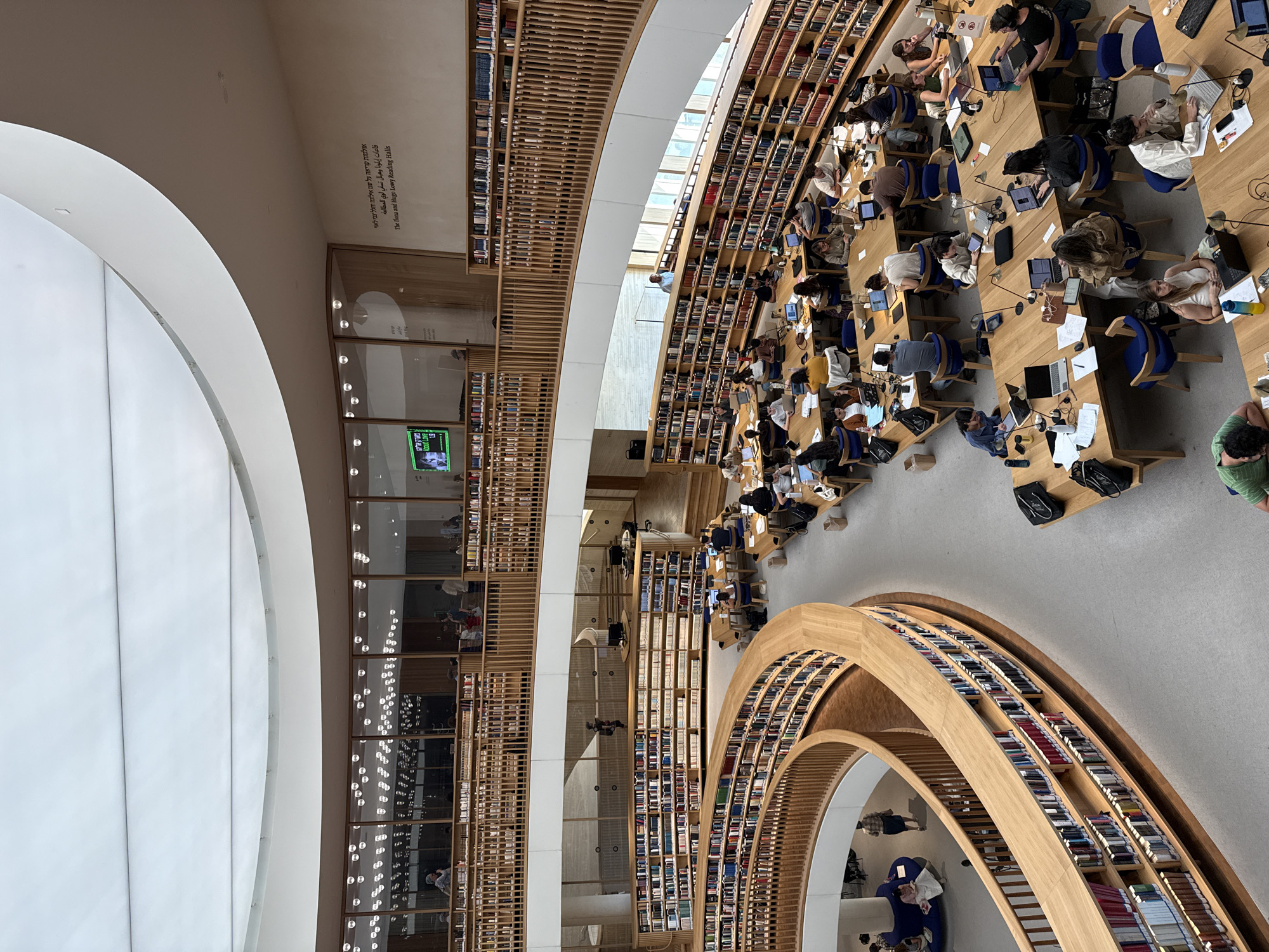 This is an interior, eye-level, wide-angle shot of a modern, multi-level library, likely the National Library of Israel in Jerusalem, Israel. The space is characterized by its expansive, curved wooden bookshelves that line the walls and balconies, filled with books. Natural light streams in from a large, overhead skylight, and a curved, white ceiling structure dominates the upper portion of the image.
Numerous individuals are scattered throughout the library, primarily seated at long wooden tables. Most are engaged in studying, working on laptops, or reading. Some are in small groups, while others are studying individually. A few people are standing or walking. The overall atmosphere is one of quiet productivity.
The library features multiple tiers with wooden railings, creating a sense of depth and architectural interest. There are numerous desks and chairs, many occupied by students and researchers. The color palette is dominated by warm wood tones, white from the ceiling and walls, and the muted colors of clothing and books. On one wall, Arabic and Hebrew text is visible, along with English text that reads "The Ilona and Hugo Lowy Reading Halls". In the upper left, a large screen displays a video. The floor appears to be a light gray, matte material.