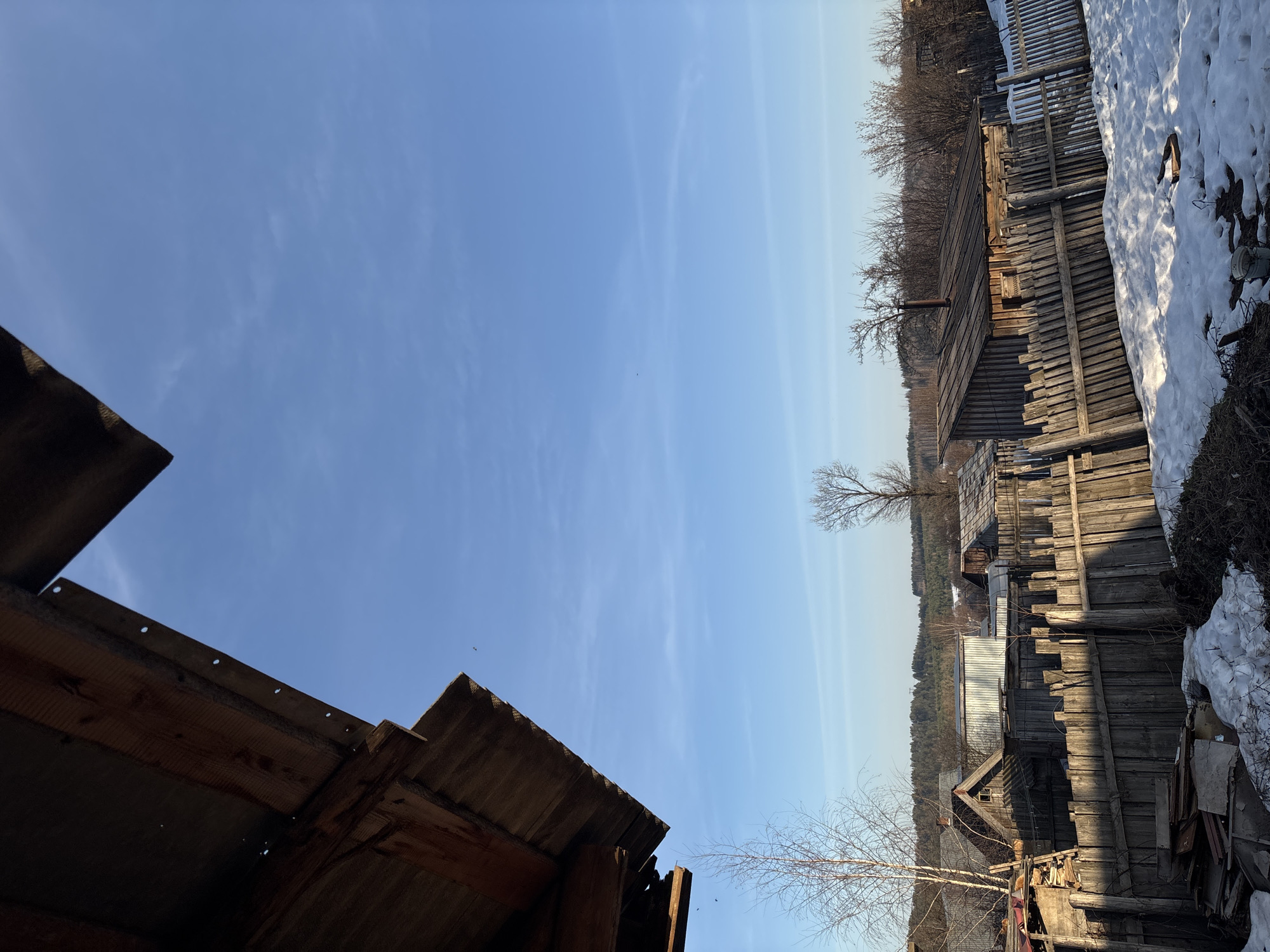 The image depicts a scene in Фоки, Russia. The perspective is angled upwards, showcasing a portion of a dark wooden structure's roof in the foreground, with a clear blue sky dominating the upper half of the frame. The background reveals a rural landscape with wooden fences, buildings, bare trees, and patches of snow, indicative of early spring.