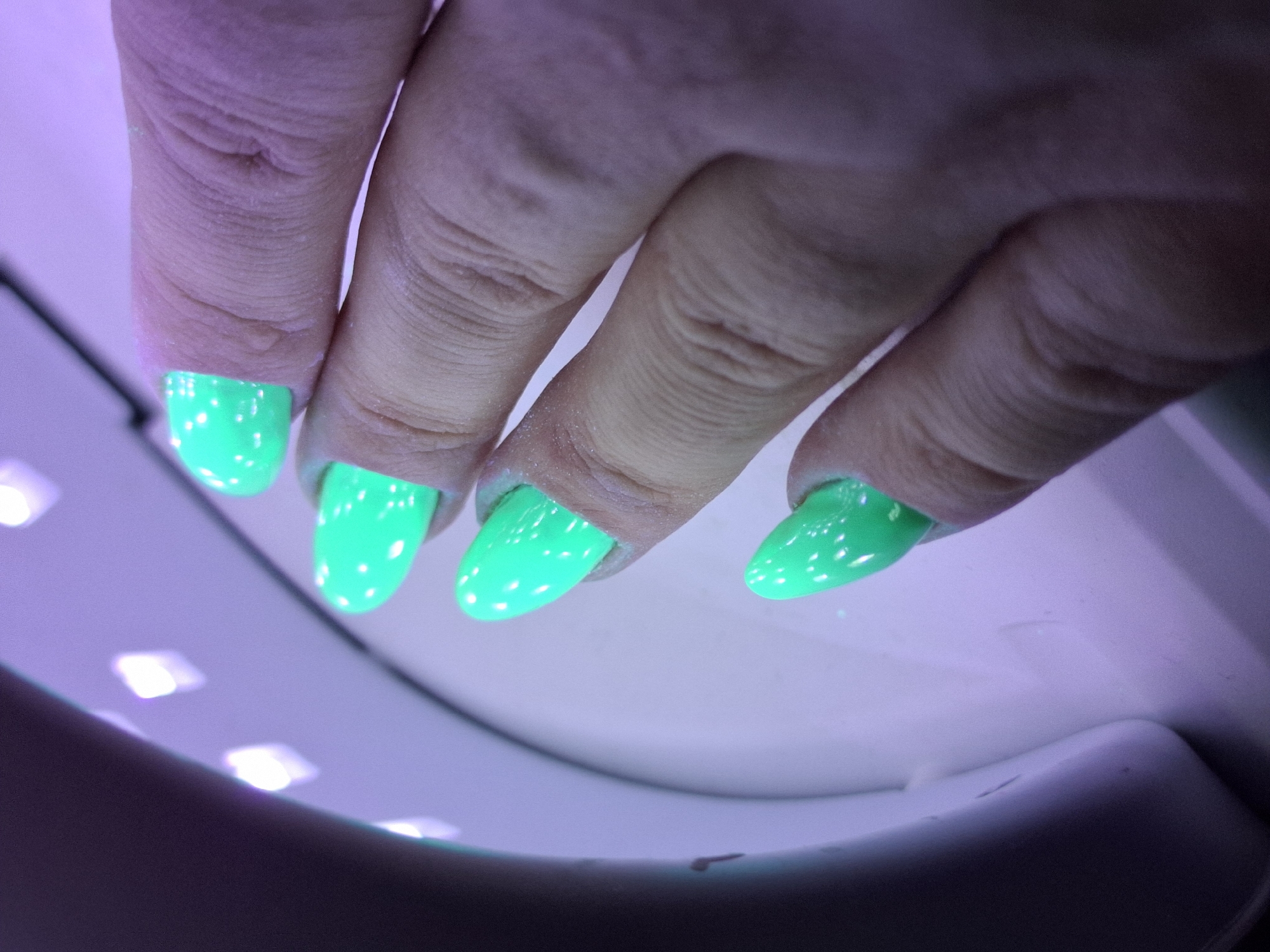 A close-up photograph depicts a human hand placed inside a nail lamp. The nails are painted in a bright green color, appearing luminous under the lamp's UV light. White spots are scattered across each fingernail. The hand is positioned above the interior of the nail lamp, which is illuminated with multiple LED lights. The color balance and lighting suggest that the photograph was taken in an indoor setting. The location of Novi Sad, Serbia, cannot be confirmed from this image alone.