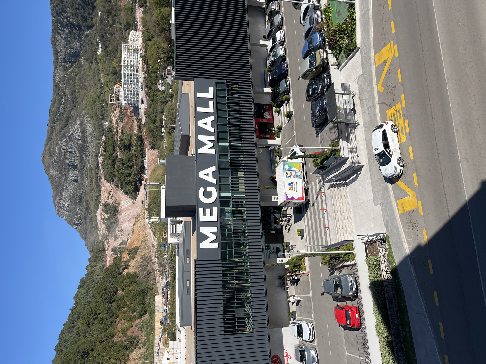 The image is an elevated view of Mega Mall in Budva, Montenegro. The mall's facade prominently displays "MEGA MALL" in large white letters. A parking lot filled with cars sits adjacent to the building, with a queue of cars in the parking lot. A white taxi is parked on the side of a road with yellow markings. A steep, rugged mountain covered in trees rises in the background, with some buildings scattered on its lower slopes. The sky is clear and blue. The overall setting suggests a commercial area in a mountainous coastal region.
