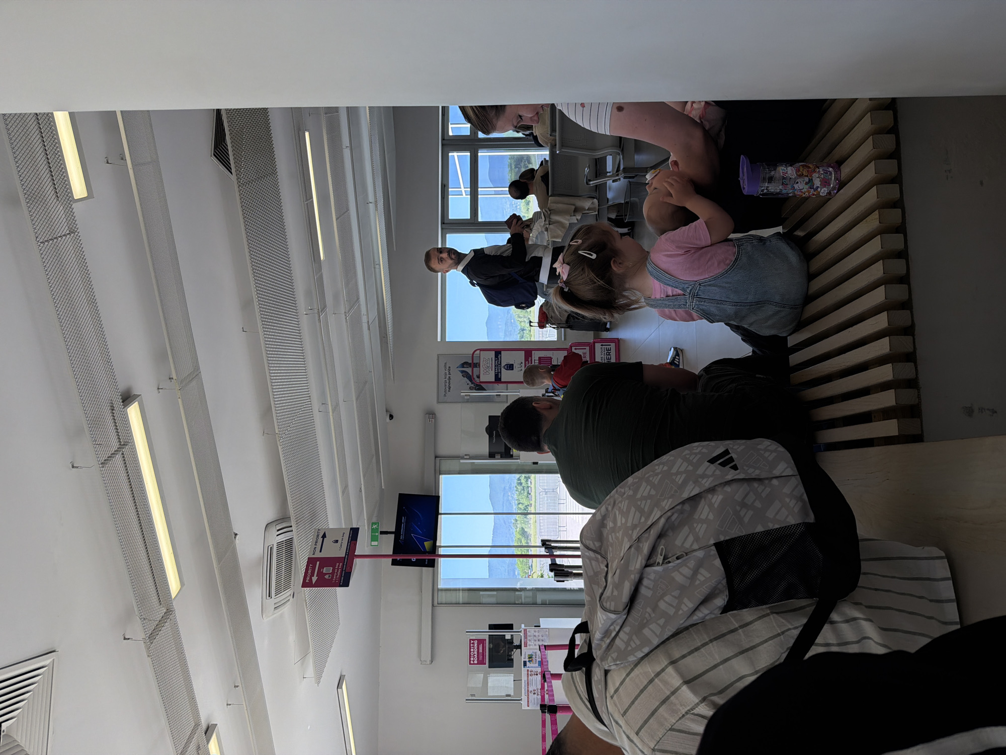 This indoor image appears to have been taken in the airport, possibly in Gornje Dubrave, Bosnia and Herzegovina. The composition reveals a group of people seated or standing, presumably waiting. Focus lies on a family of four, including a young girl in denim overalls and a pink shirt, gazing upward towards her mother. An adult male, who may be the girl's father, wears a green shirt and sits nearby. Another man wearing a dark jacket stands near the window, in which hills are visible. Luggage and bags are strewn around, including a backpack. The environment is illuminated by overhead lights, and the scene is typical of an airport waiting area. The decor includes advertising and signs.