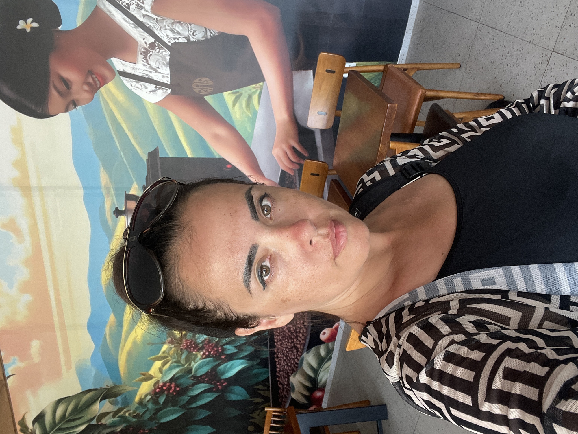 A woman with dark hair and sunglasses perched on her head is captured in a selfie with a mural in the background. The mural depicts a woman in traditional attire with a flower in her hair, smiling and appearing to be working with coffee beans. The background also features a landscape with rolling hills and a sky in hues of blue and orange, suggesting a daytime setting. In the foreground, the woman is wearing a black top with a black and white patterned sheer overlayer. Her expression is neutral, with her eyes open and looking towards the camera. Visible in the background are wooden chairs. The overall scene appears to be indoors, possibly in a cafe or restaurant with decorative wall art. The image is oriented sideways, with the subjects appearing rotated.