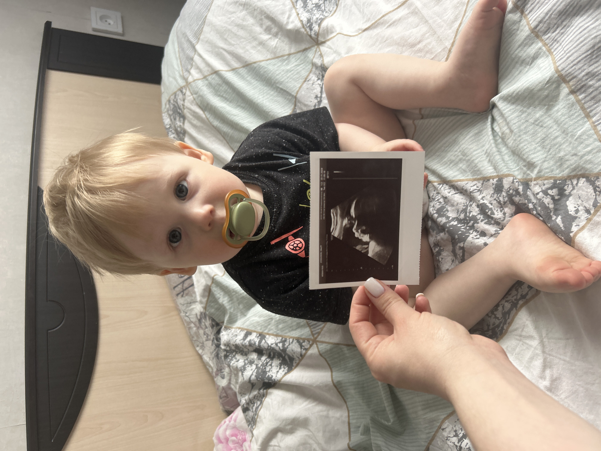 An indoor, overhead shot captures a blonde toddler with bright blue eyes and a green pacifier, lying on a patterned duvet on the floor in what appears to be a home in Chaikovsky, Russia. The child wears a black t-shirt with a small graphic, possibly an anchor or fishing hook design, and looks directly at the camera with a curious expression.
In the foreground, a woman's hand, with neatly manicured white nails, holds up a black and white ultrasound printout. The ultrasound image clearly displays the profile of a fetus. Visible text on the printout includes medical abbreviations and numbers such as "OB", "PW", "CRL", and "6.24cm". A date "05.10.2023" and gestational age "12w 2d" are also discernible, indicating an early pregnancy.
The child is positioned on a duvet with a geometric pattern in shades of grey, white, and muted green, transitioning into a floral pattern with pink details towards the bottom left. The background features light-colored wooden flooring, a dark wooden furniture piece (possibly a headboard or paneling), and a light-colored wall with a white electrical socket visible near the top left corner. The bright, even lighting suggests it is daytime. The scene evokes a personal family moment, possibly an announcement of a new pregnancy.