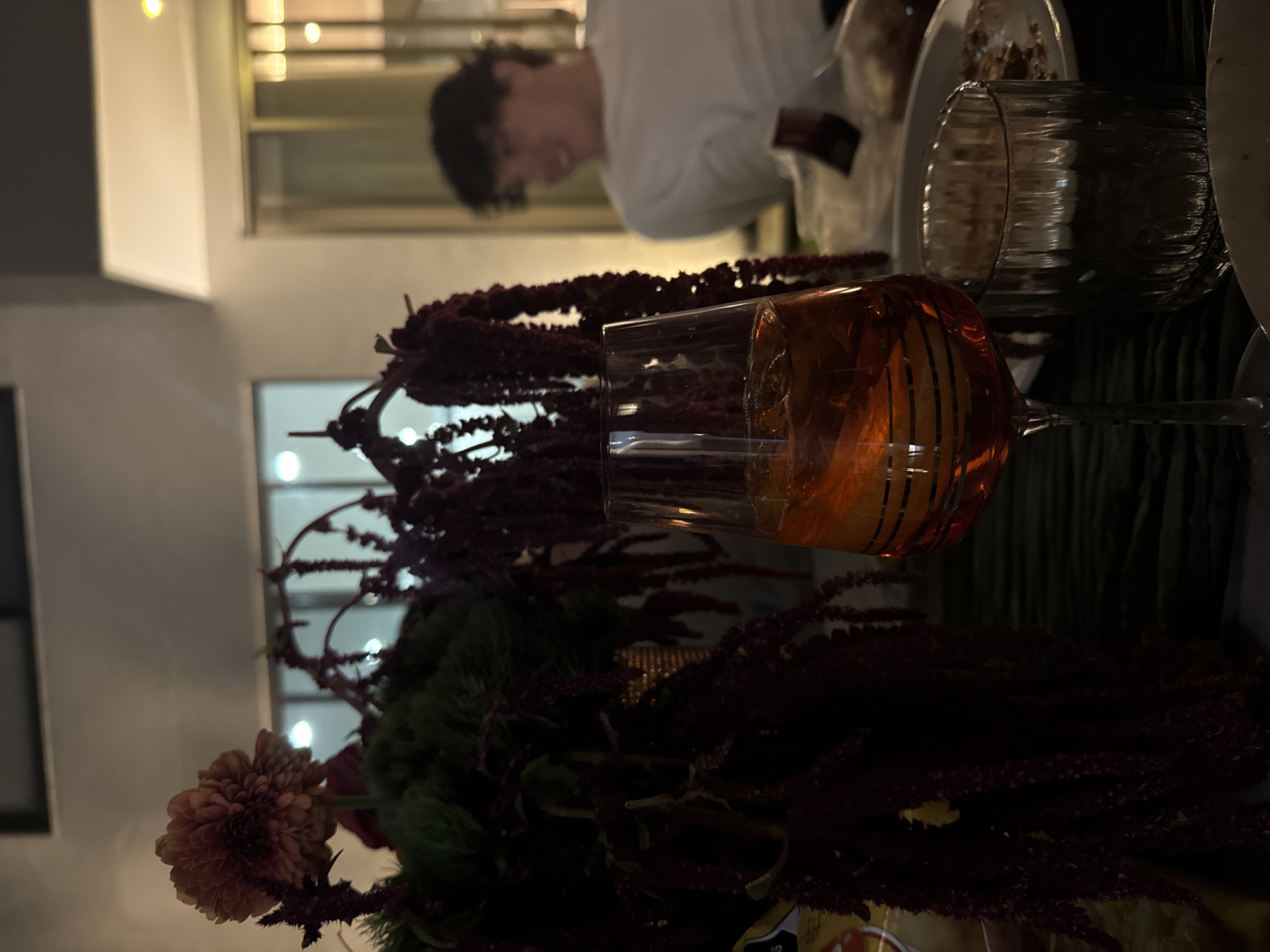 The scene depicts an evening gathering, likely on a patio or terrace, in Puebla, Mexico. In the foreground, a dark, textured surface, possibly a tablecloth, supports several items. A prominent floral arrangement occupies the left half, featuring a large, reddish-orange chrysanthemum-like bloom at the top, dark green foliage, and long, cascading strands of dark red amaranthus. A tall, stemmed glass filled with an orange cocktail and ice cubes stands centrally, displaying horizontal dark stripes as a design element. To its right, a shorter, clear, fluted glass is visible. Partially seen white plates with food remnants and a yellow and white bag (possibly snacks) are also on the table. In the midground, behind the glasses and flowers, a young male with dark, curly hair and a white t-shirt is seated, his face turned left and appearing to smile or laugh; his features are blurred due to shallow depth of field. The background consists of light-colored architectural elements, likely walls and large windows or glass doors, through which bright interior lights are visible, casting reflections. The overall illumination is artificial and dim.