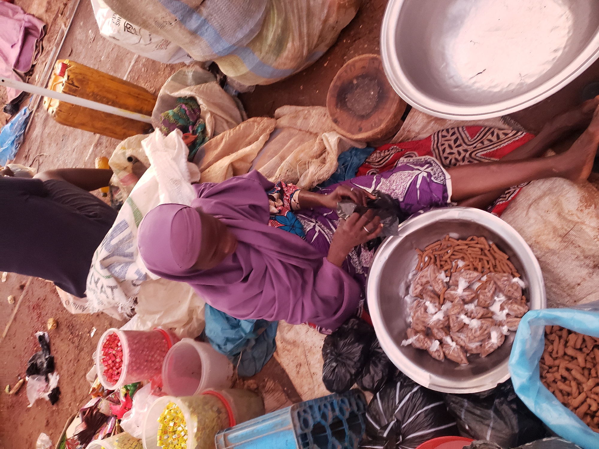 In Kakuri Gwari, Nigeria, a person wearing a purple hijab and a patterned dress sits on the ground next to a variety of goods. They are handling a small, dark object. Surrounding them are several large sacks, plastic containers filled with colorful items, a large yellow jug, black trash bags, and a metal bowl containing what appears to be food items packaged in plastic. A wooden cutting board is visible to the right of the bowl. The ground is covered with reddish-brown dirt, and debris is scattered around the area. Another person, partially visible in the background, is wearing a dark shirt. The scene appears to be an outdoor market or storage area.