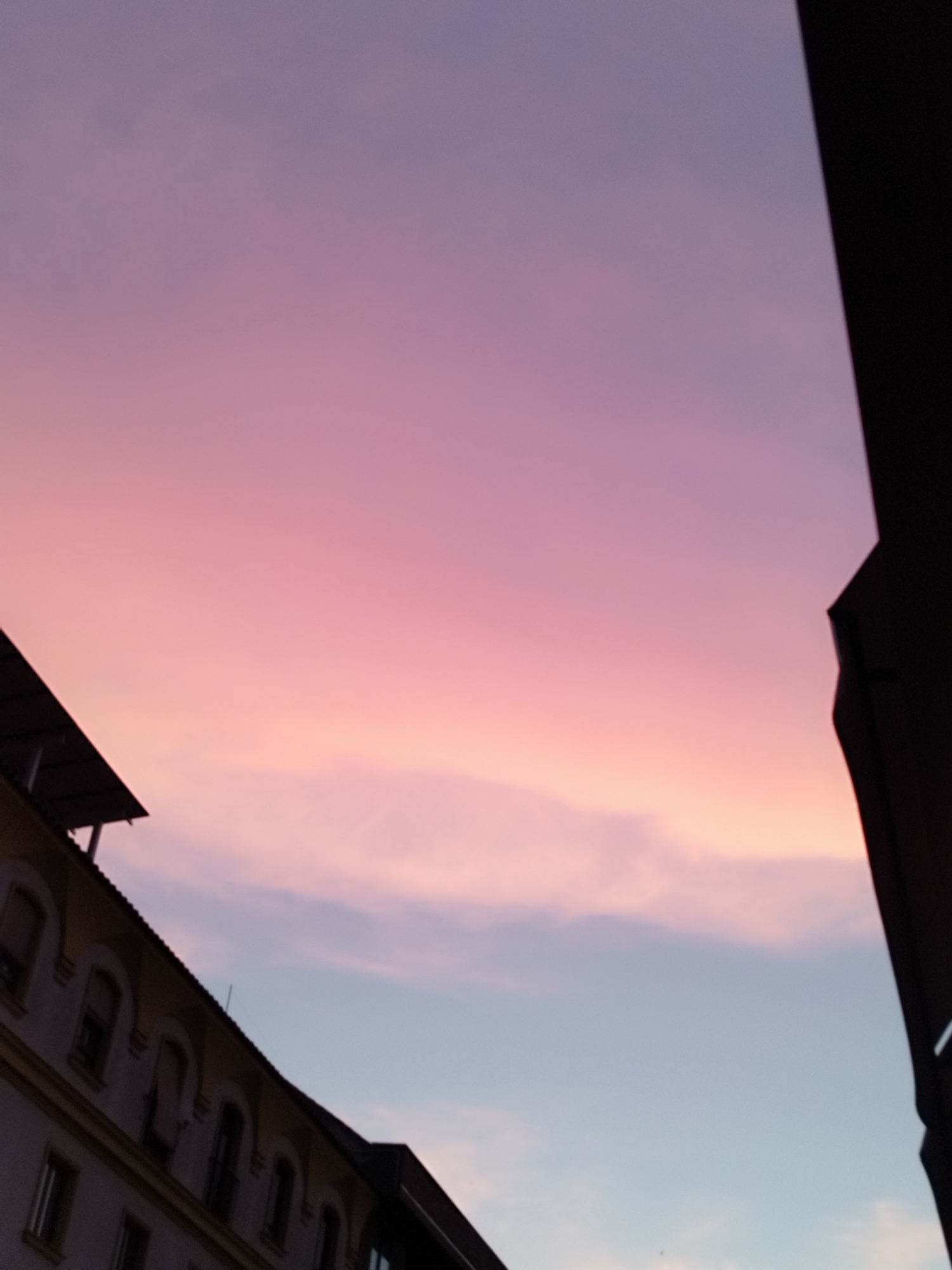The image depicts a twilight sky with hues of pink and purple transitioning to a pale blue. A portion of a building, likely in the city of Jerez, Spain, is visible in the lower left quadrant. The building has a mansard roof and multiple arched windows. A dark, angular structure, possibly another building or architectural element, occupies the right side of the frame, obscuring part of the sky. The scene is static, with no visible people or ongoing actions. The environment appears to be urban, characterized by the presence of the building against the backdrop of the evening sky.