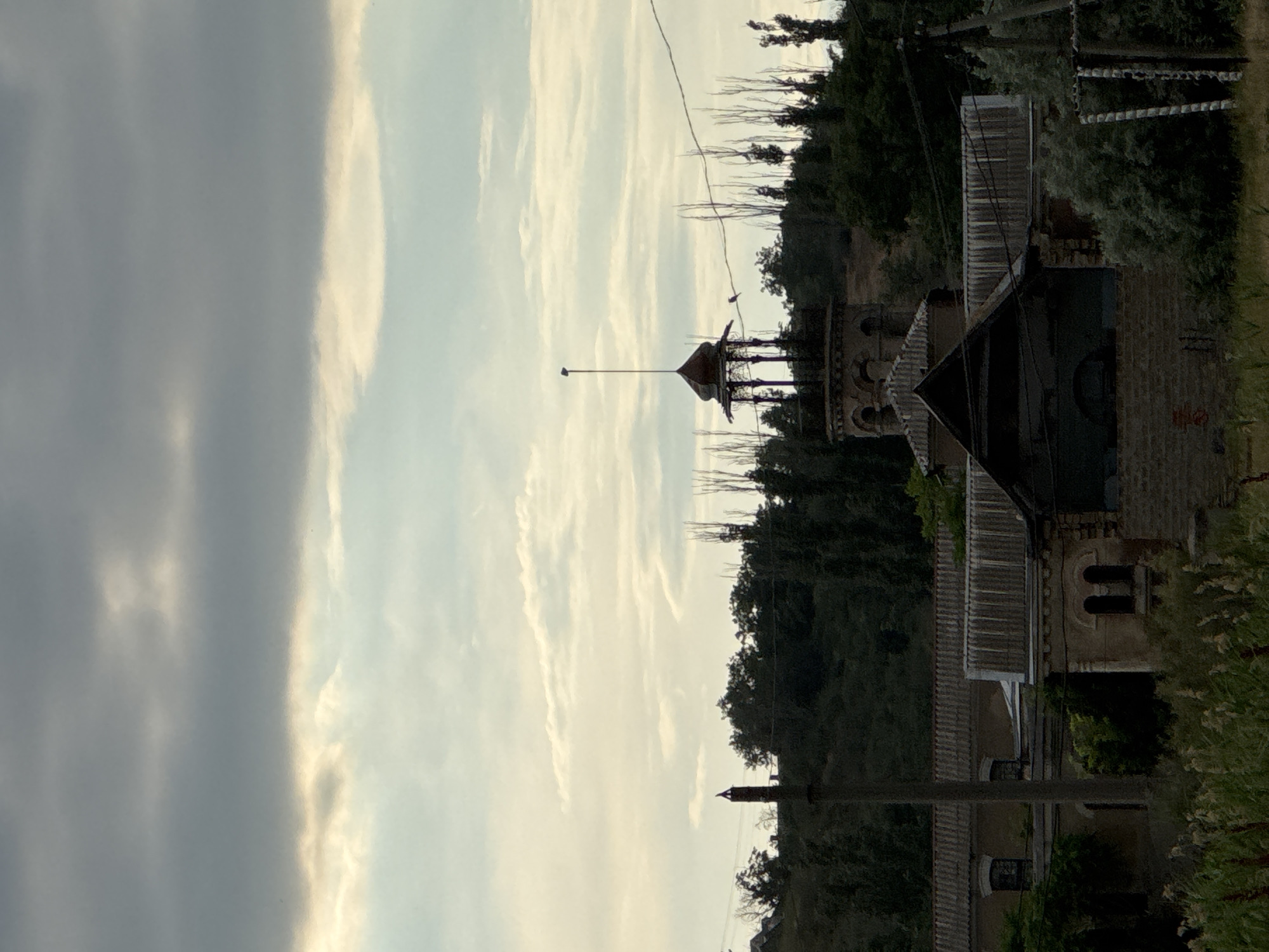 The image depicts a building with a distinctive turret against a backdrop of trees and a cloudy sky. The building appears to be made of brick and has a weathered, old-fashioned architectural style, possibly a historical structure or an older residence. The turret is topped with a pointed roof and has a rod extending upwards. A prominent power line runs horizontally across the upper portion of the scene, with two small birds perched on it. The surrounding area is densely covered with dark green trees, suggesting a rural or semi-rural setting. The sky is a mix of dark gray clouds on the left and lighter, wispy clouds on the right, indicating possibly overcast or a transition in weather. The lighting suggests it is either early morning or late afternoon. The overall mood of the image is somewhat somber or mysterious, due to the dark tones and the overcast sky. No visible text is present in the image.