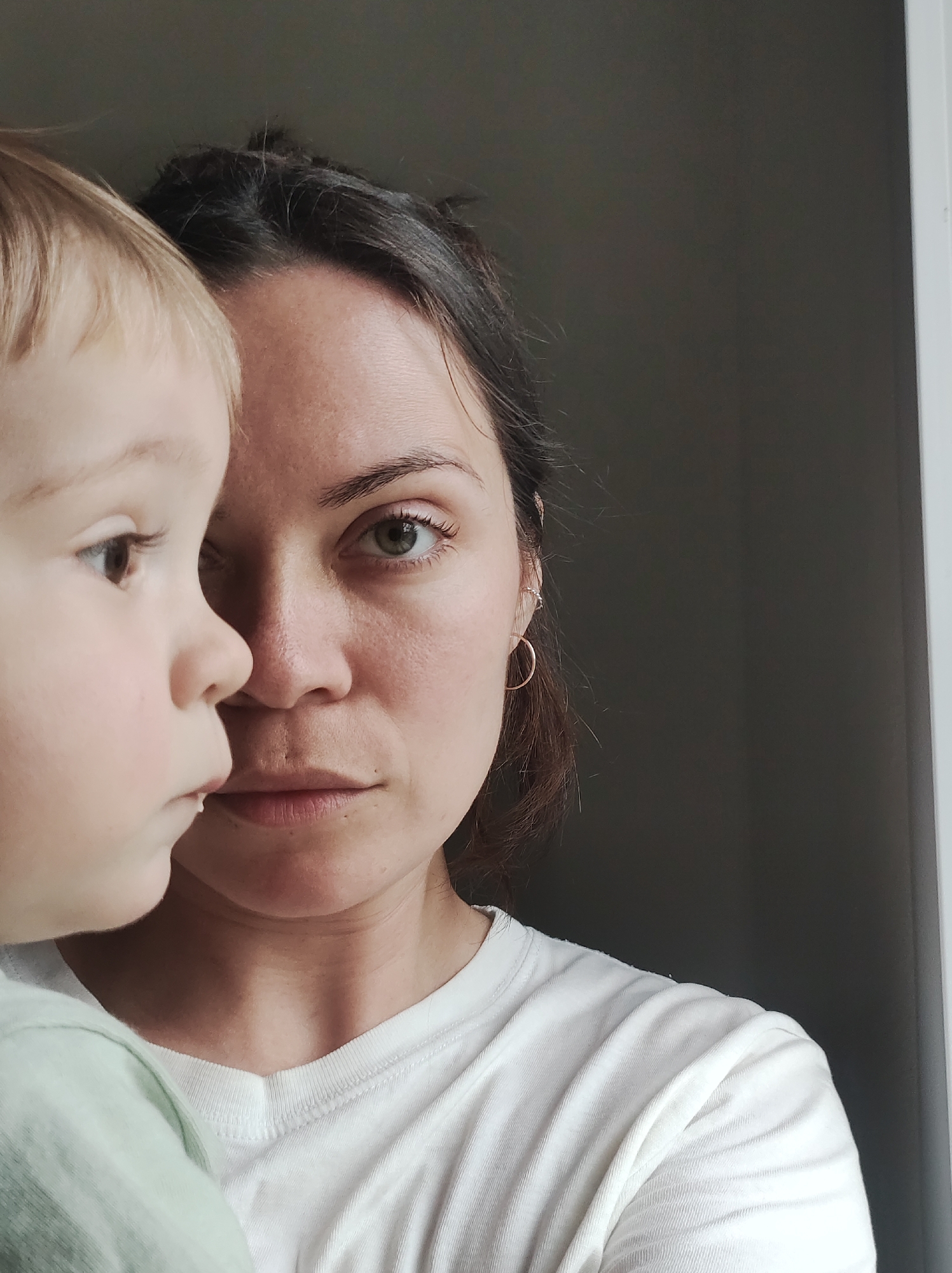A close-up shot shows a woman and a young child positioned closely together. The child, with blond hair, is positioned to the left, facing away from the camera, with only their profile visible. The woman, with dark hair pulled back, faces the camera directly, her gaze intense. She wears a white t-shirt and a small, gold hoop earring. The background is a plain, dark wall.