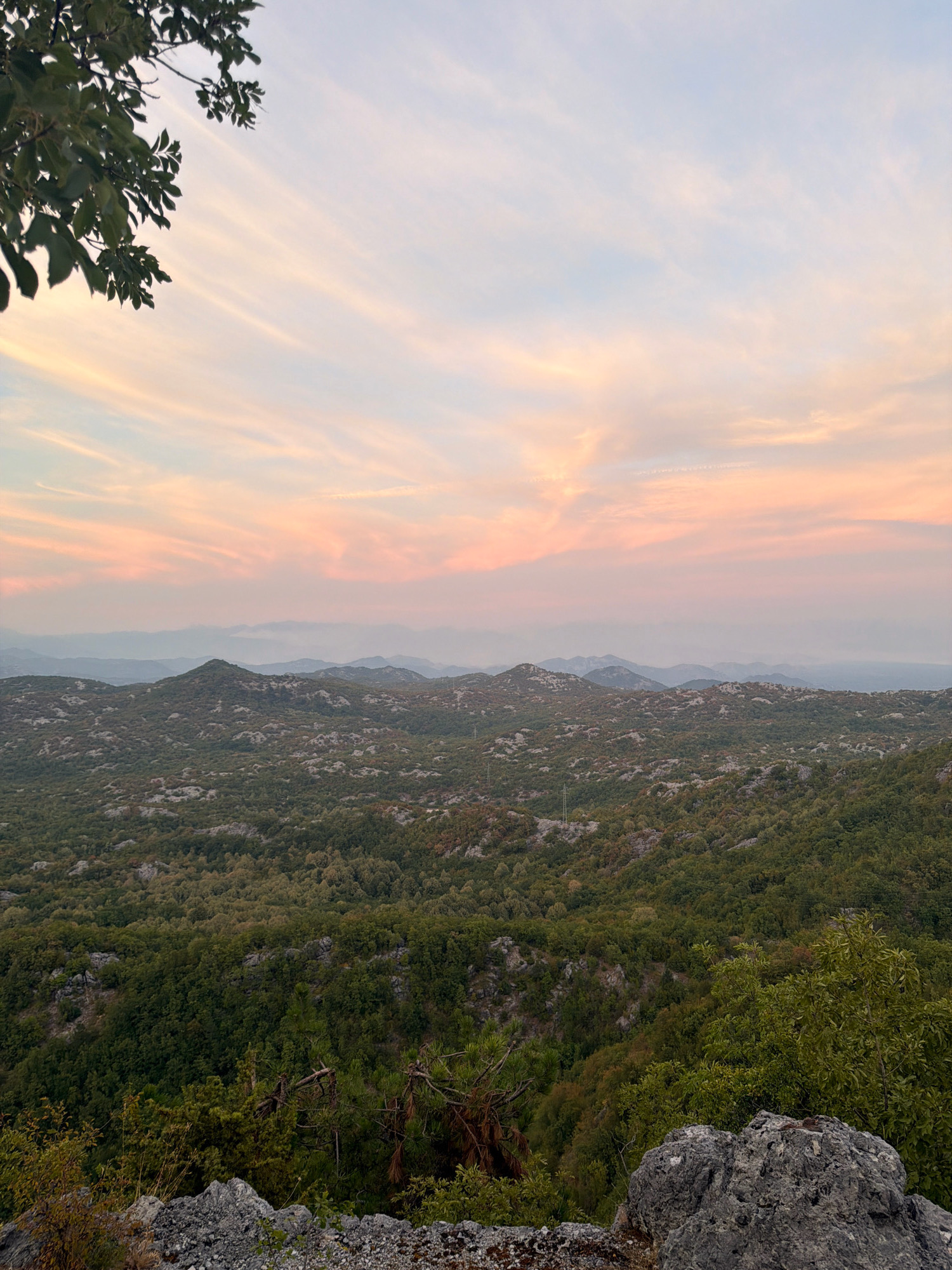 The image captures a panoramic view of a mountainous landscape at dusk, likely from a high vantage point in or near Boguti, Montenegro. The sky is painted with soft hues of pink, orange, and pale blue, with delicate clouds stretching across the horizon. In the foreground, a rocky outcrop and some foliage frame the scene. The mid-ground is dominated by rolling hills covered in dense, green vegetation. Further in the distance, successive layers of mountains recede into a hazy, muted landscape, indicating a significant expanse. The overall atmosphere is serene and natural.