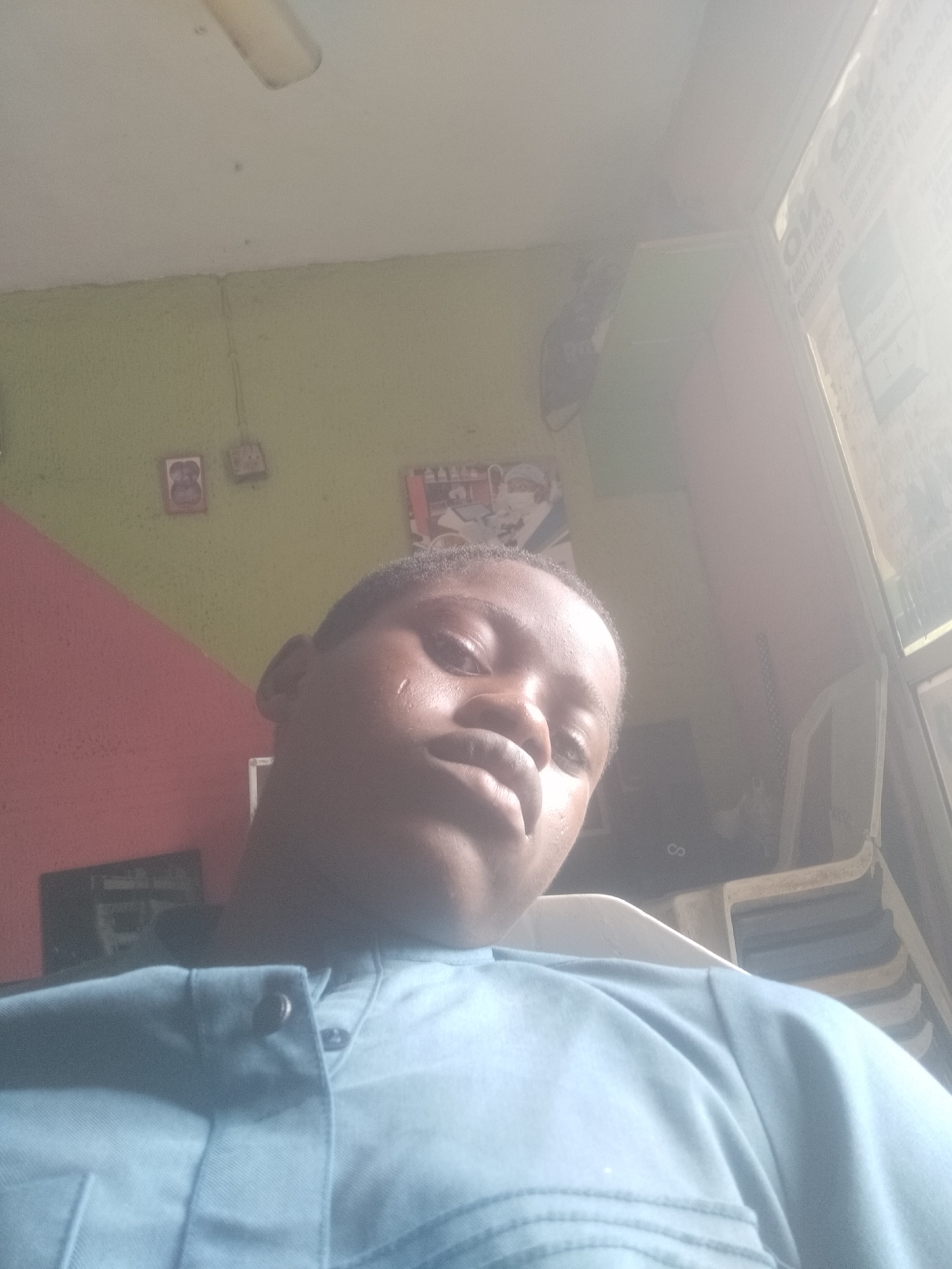 A low-angle indoor shot depicts a person with dark skin wearing a light blue collared shirt. The person is facing the camera with a slight upward gaze. A small droplet is visible on their cheek.
The background consists of a light green wall with hints of red near the left edge. Several small framed images or objects are mounted on the wall. A window is located in the upper right corner of the frame, letting in a significant amount of light. In the background, chairs with white plastic frames can be partially seen.
The location is suggested to be Shangisha, Nigeria.