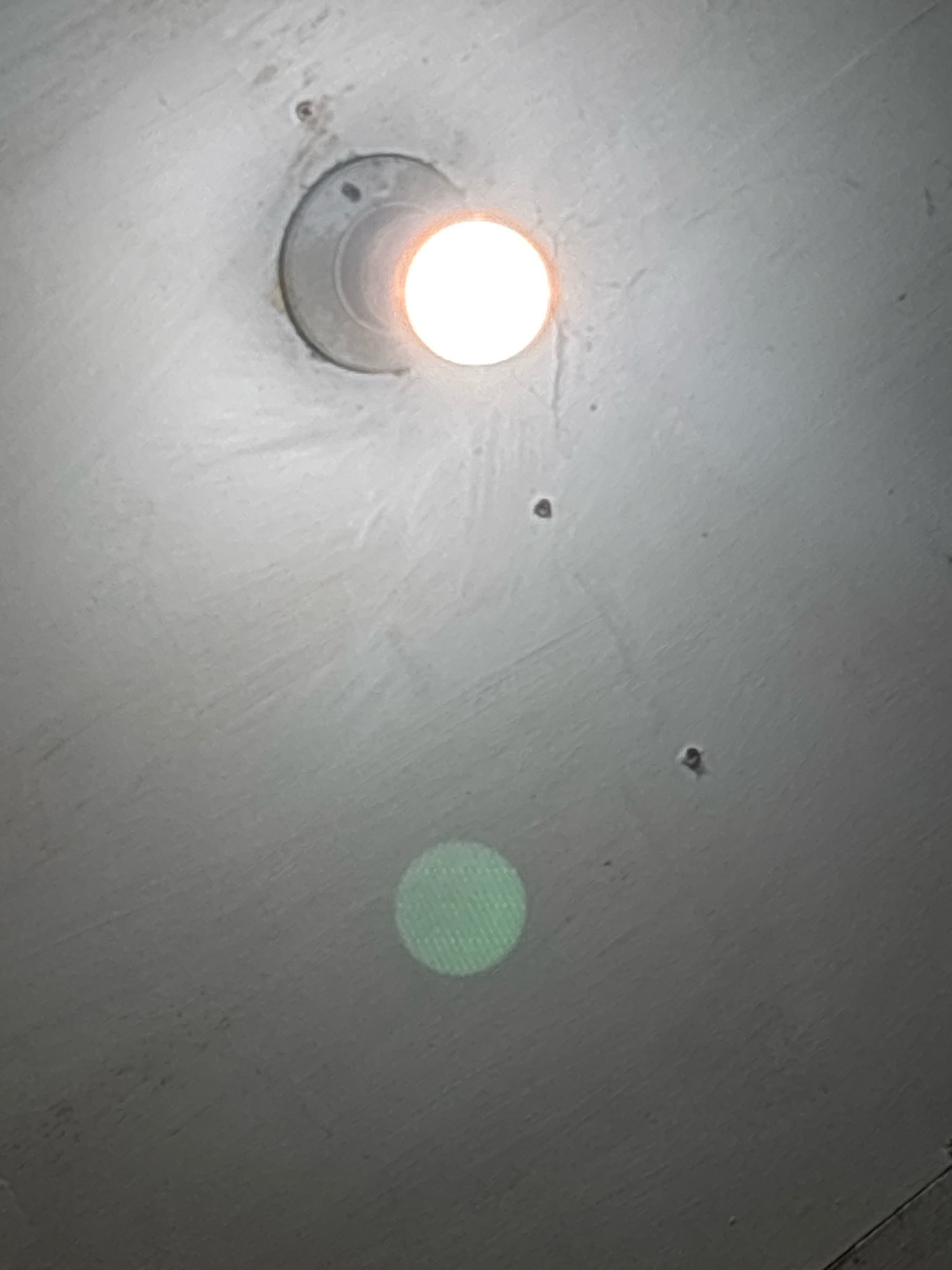 The image shows a ceiling with a light fixture and a bright light emanating from it. There is also a faint green circular reflection on the ceiling. The ceiling appears to be made of concrete and has some minor imperfections and markings. The image is taken from a low angle, looking upwards towards the ceiling. The location of this scene in North Jakarta, Indonesia is not discernible from the provided image.