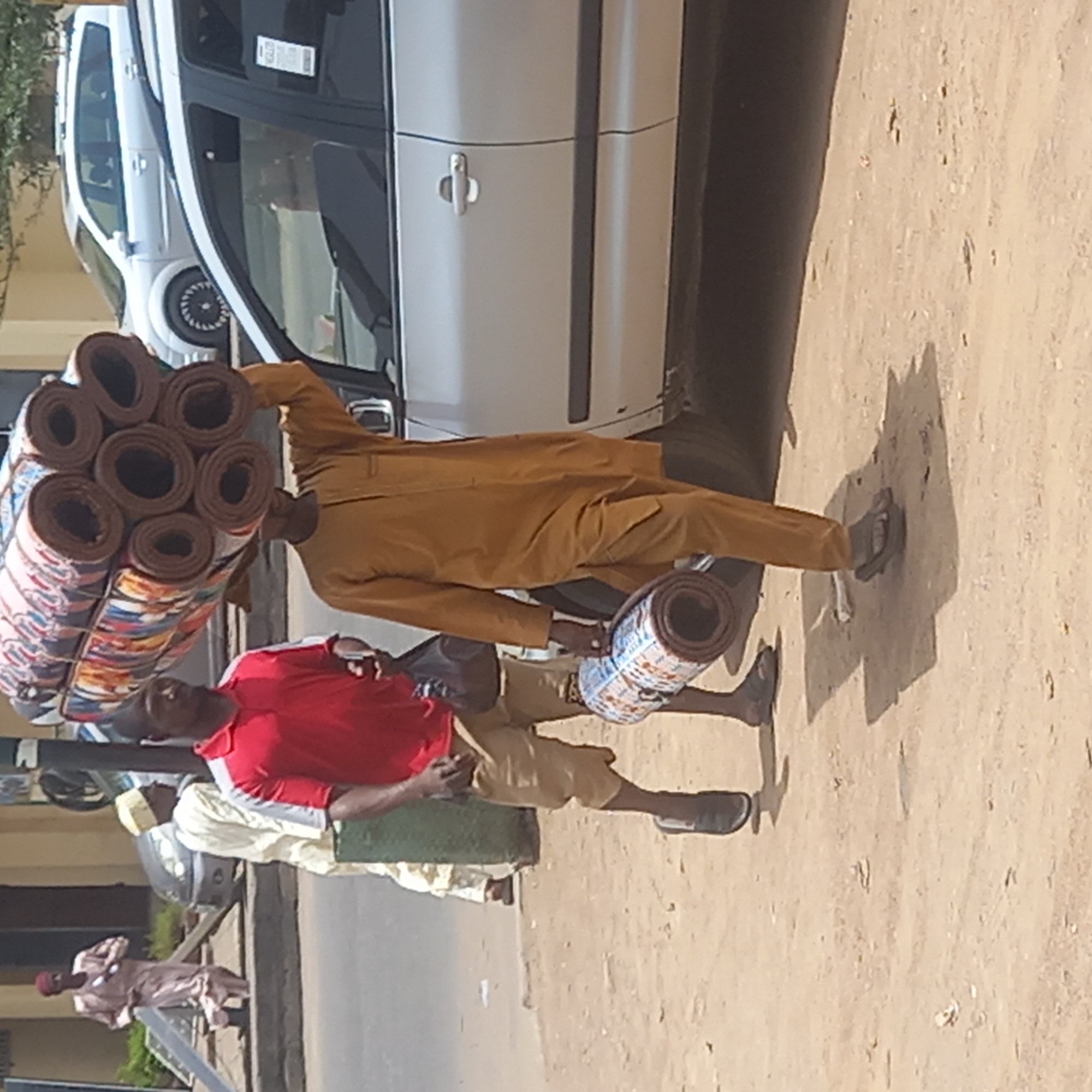 The image captures a sunny street scene in Dadi Riba, Nigeria, depicting daily life and commerce. In the foreground, a man dressed in a light brown traditional outfit (possibly an agbada) carries a tall stack of colorful, rolled mats on his head, with another rolled mat held in his left hand. The mats are patterned and appear to be textiles or rugs. He is walking on a light brown, sandy-dirt surface.
Beside him, another man wears a red short-sleeved shirt and light brown shorts, standing near the first man. Both men cast strong shadows on the ground, indicating bright daylight, likely midday or early afternoon.
In the background, a white car is parked on the side of the road, partially visible with its windows and a door handle. Behind it, part of another white vehicle, possibly a larger van or bus, can be seen. Further to the left, on a slightly elevated pavement, another person in a light pink traditional outfit walks. The ground transitions from sandy dirt to an asphalt road further in the background. The overall scene suggests a busy street or market area where goods are being transported or sold. No legible text is visible on any objects.