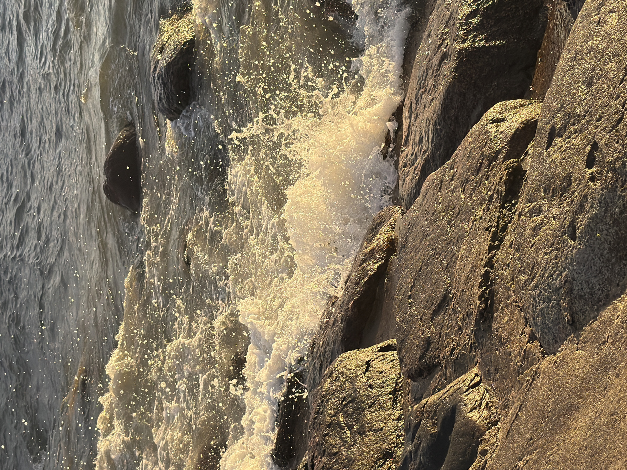 The image depicts a close-up view of waves crashing against a rocky shoreline. The water is agitated, with white foam and spray being thrown up as the waves hit the rocks. Sunlight glints off the water's surface, creating a sparkling effect. The rocks in the foreground are dark and wet, with some appearing to be covered in a thin layer of water. The background shows more of the choppy water extending towards the horizon. There are no people or man-made structures visible in the image. The scene suggests a natural coastal environment. Based on the provided context, this location is in the city of Tallinn, Estonia.