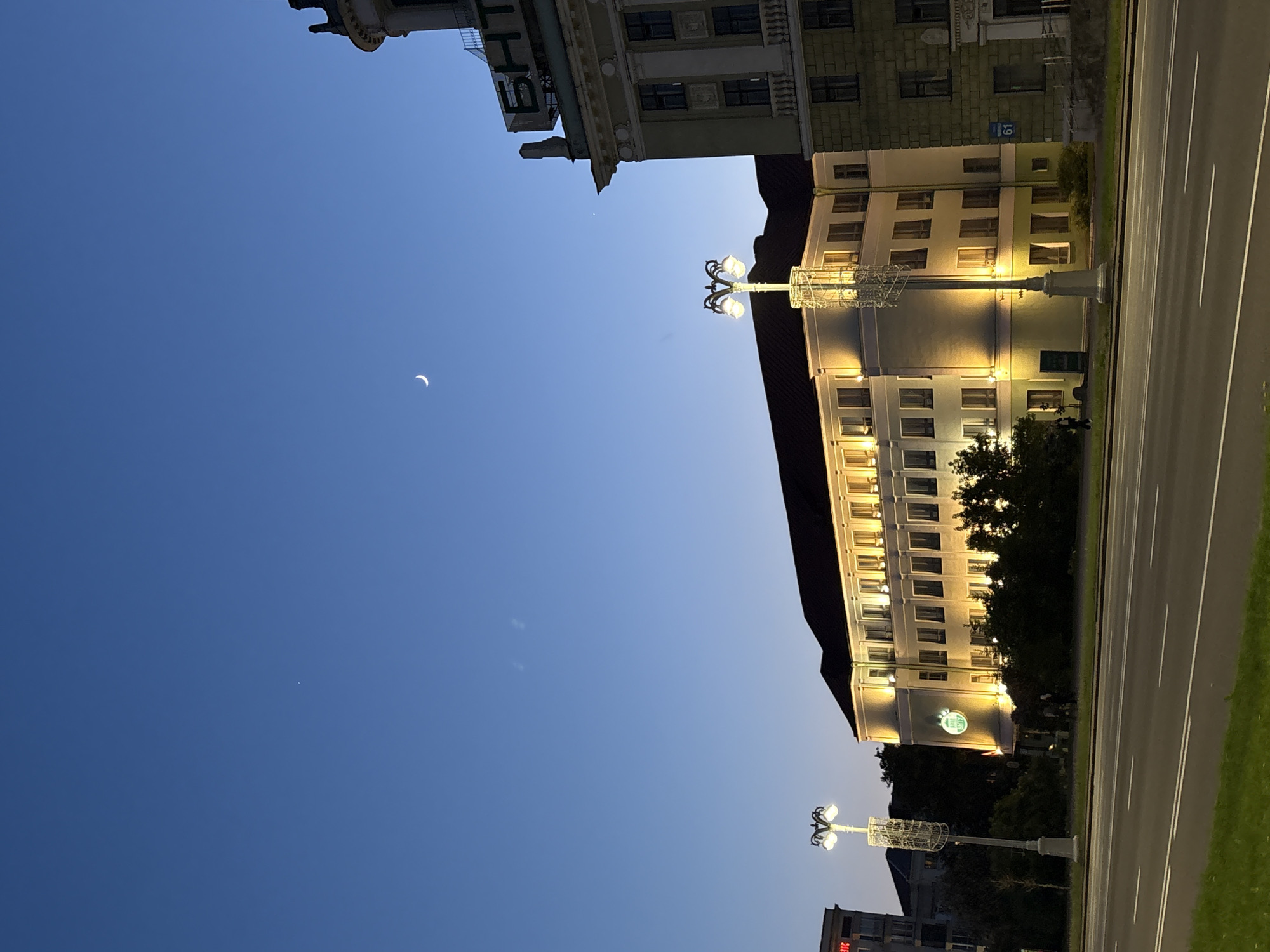 The photo shows a street in Minsk, Belarus at dusk. A road with white lines runs vertically on the right side. The facade of a tall, light-colored building is on the right, illuminated by warm-toned street lights. The night sky is a gradient of blue, darkening towards the top, with a crescent moon visible. A second building is visible at the top. The photo is tilted.