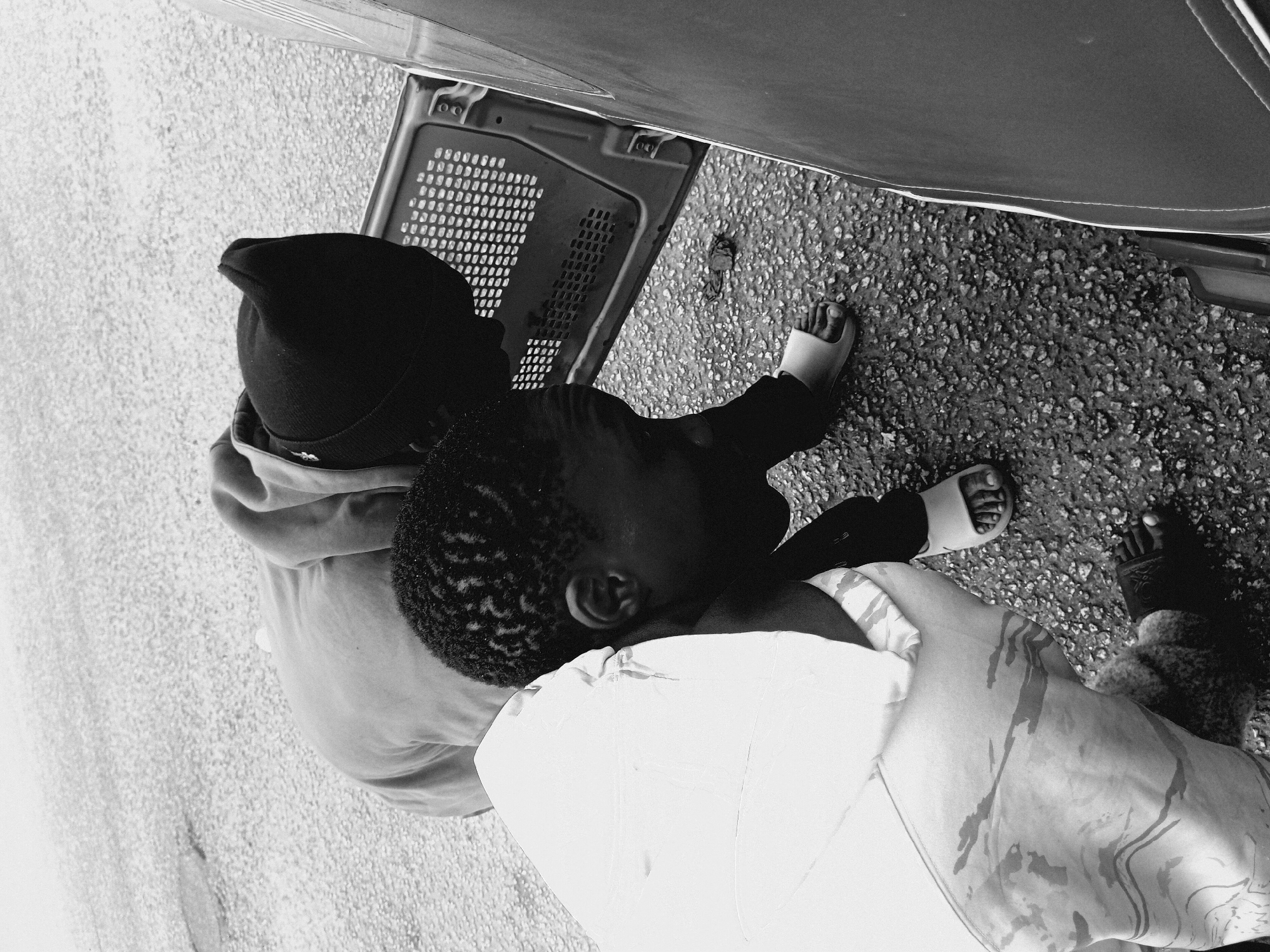 The image, in black and white, depicts two individuals standing near a vehicle. The first individual is wearing a dark-colored beanie, a light-colored jacket, and dark pants with white sandals. Their head is near a metal grate. The second individual is wearing dark pants and one dark sandal with a light colored blanket. The ground appears to be asphalt or concrete. It is not possible to confirm the location within Bukuru, Nigeria, without additional information.