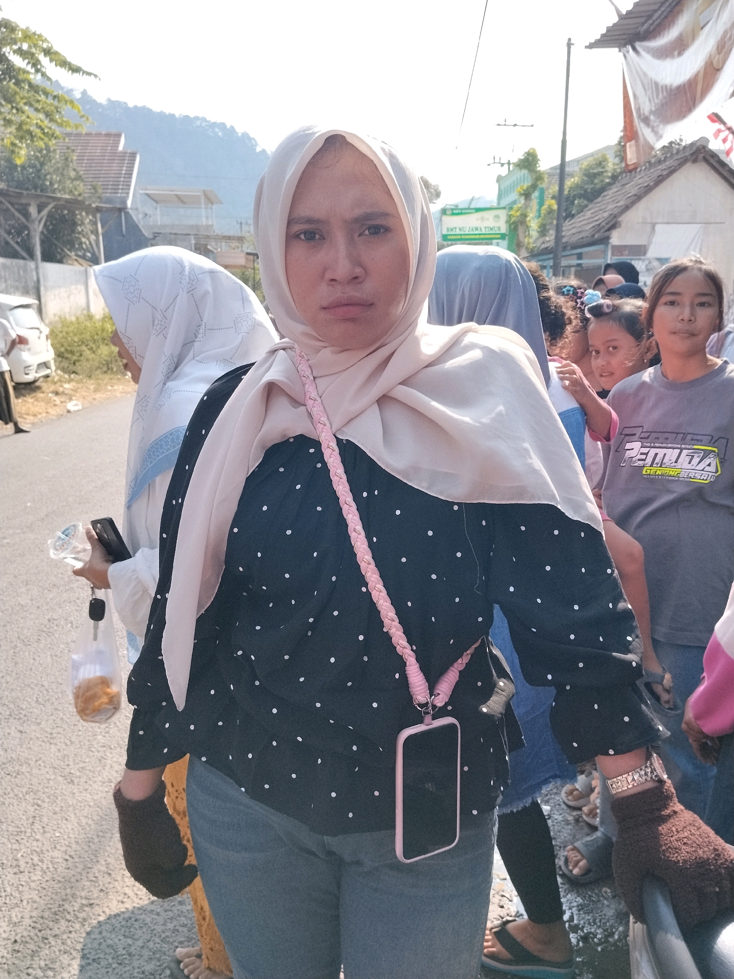 In Tlogosari, Indonesia, a medium shot captures a group of people standing on an asphalt road under bright daylight. The central subject is a woman wearing a light beige hijab and a black, polka-dotted shirt. She has a phone case with a pink braided strap around her neck, a brown glove on her right hand, and a silver-toned bracelet on her left wrist. To her left, another woman in a white hijab is visible, and behind her, several children and adults are gathered. Some of the children have their hair styled with colorful accessories. The background features houses, trees, and a distant mountainous landscape. A sign with Indonesian text is mounted on a pole. The overall scene appears to be an outdoor gathering or event.
